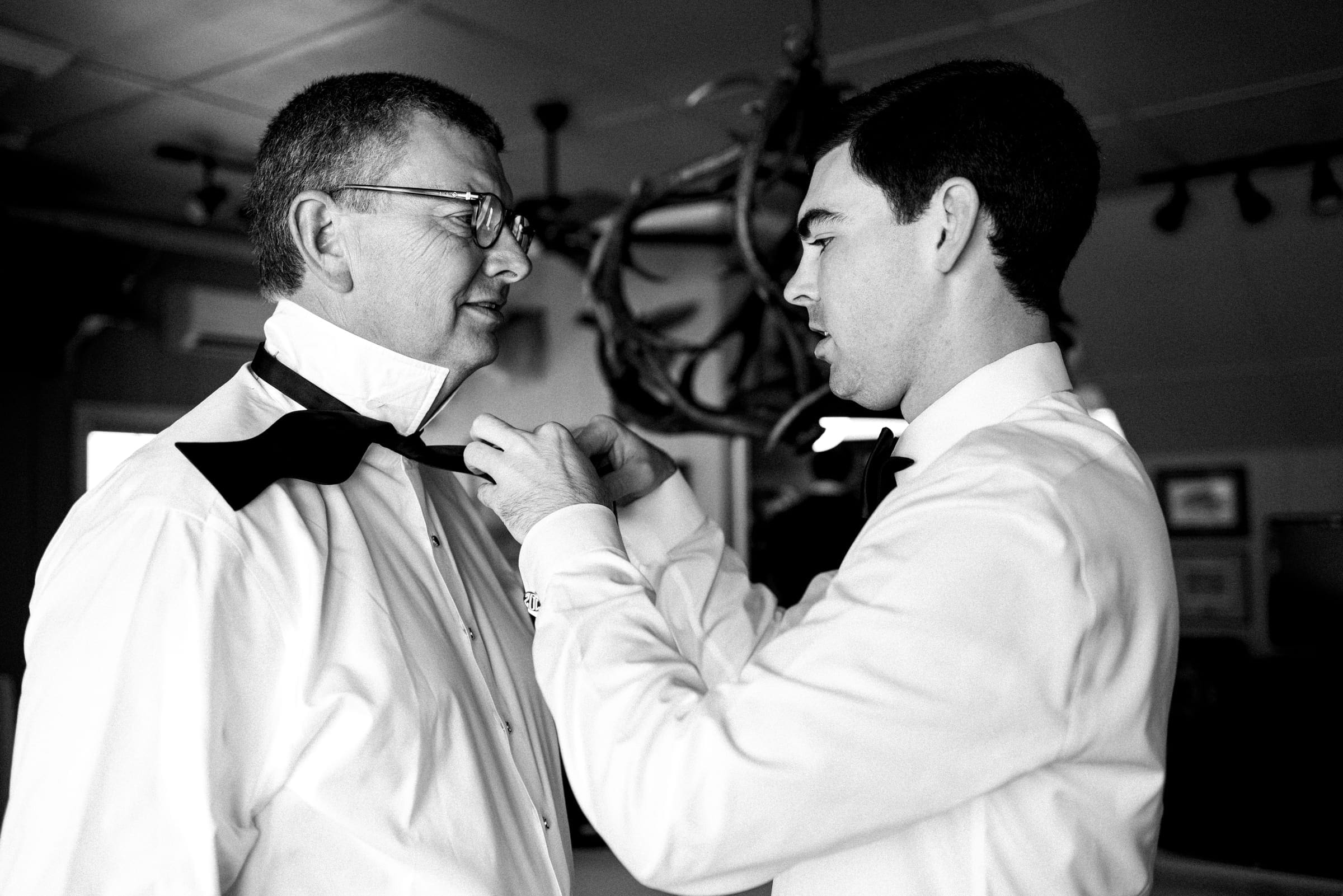 dunn wedding photography - reaves & zach In a dimly lit room, a young man skillfully adjusts an older gentleman's bow tie, both clad in formal white shirts, as they prepare for a special occasion. The elegant setting is perfect for Dunn wedding photography, with a decorative light fixture casting a warm glow in the background.