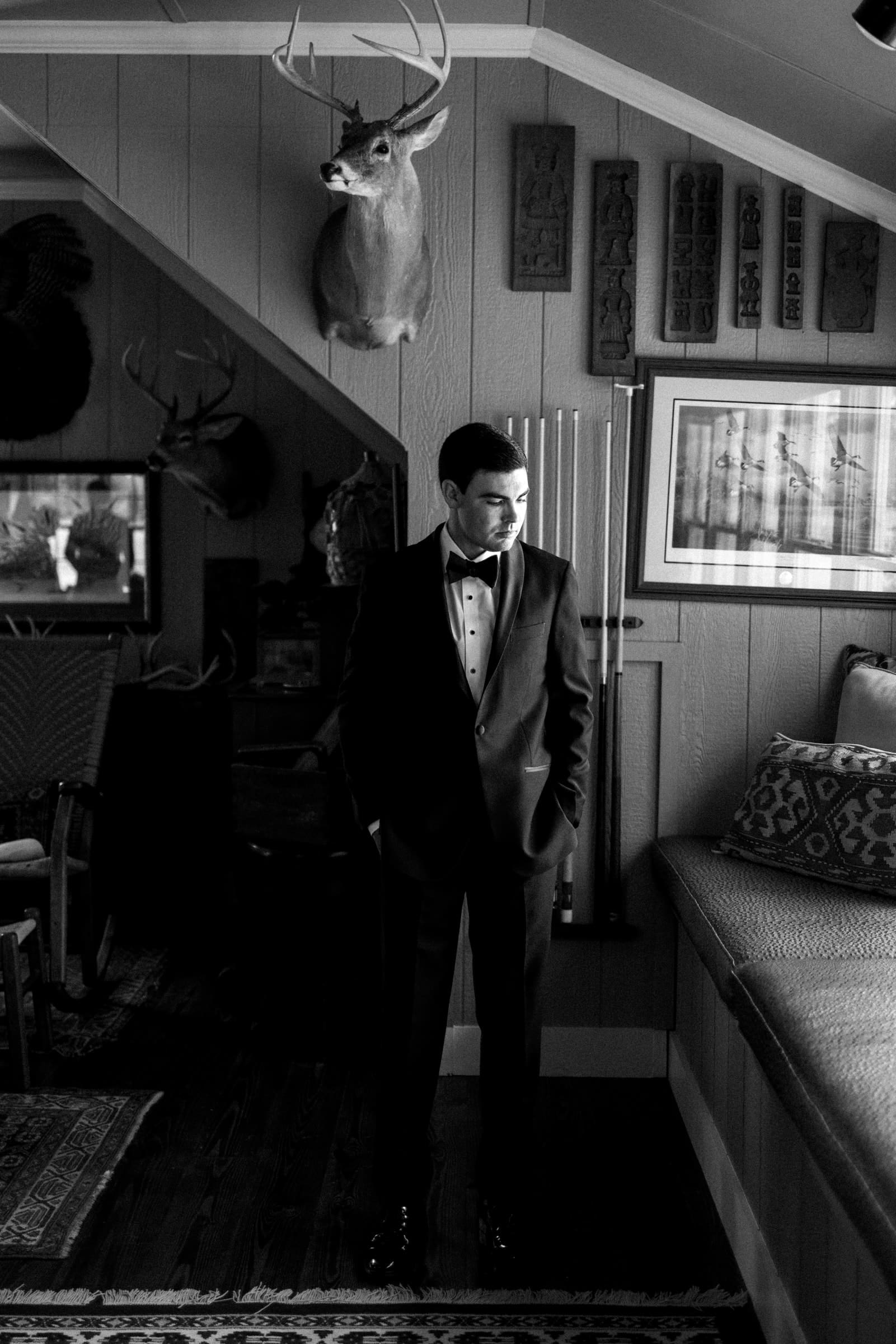 dunn wedding photography - reaves & zach A man in a tuxedo stands in a dimly lit room, reminiscent of Dunn wedding photography, decorated with hunting trophies and art. The room features wooden paneling, a deer head mounted on the wall, and framed artwork. The atmosphere is sophisticated and rustic.