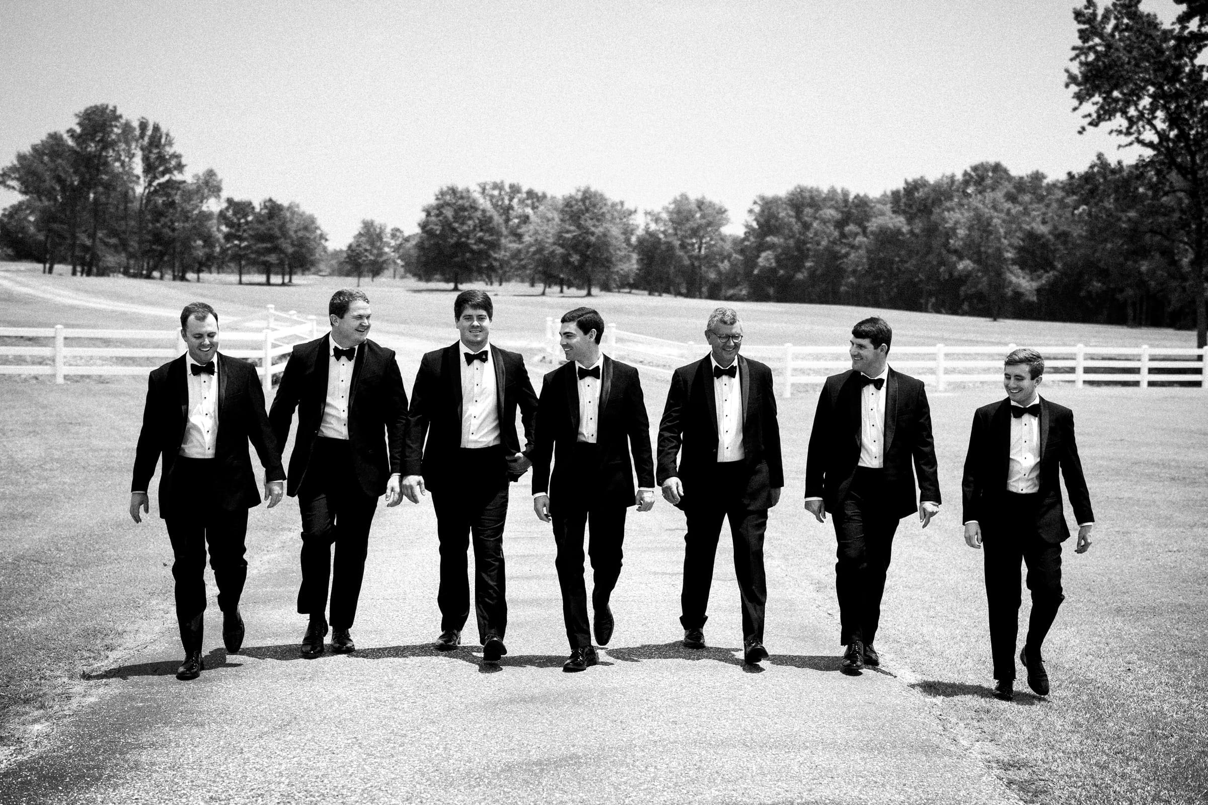 dunn wedding photography - reaves & zach A group of seven men in tuxedos walks confidently on a paved road, silhouetted against trees and a white fence under a clear sky. Captured in black and white by Dunn Wedding Photography, the image exudes a timeless, classic elegance.