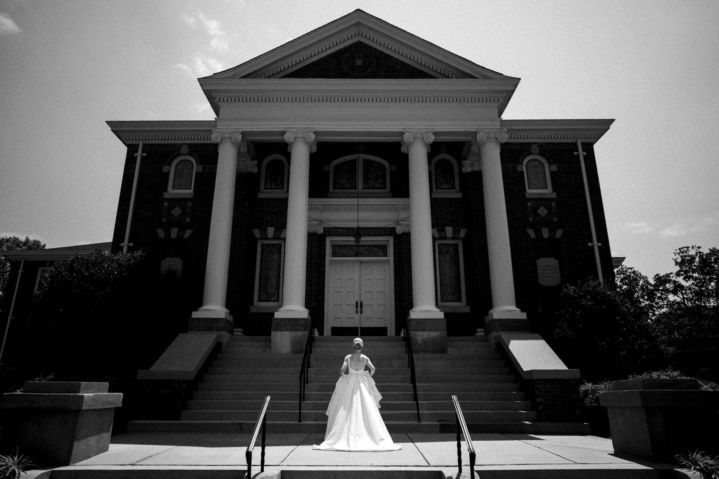 dunn wedding photography - reaves & zach A bride in a white dress stands on the steps of a large, traditional building with tall columns and ornate architecture. Captured by Dunn wedding photography, the black and white scene highlights the elegant contrast and the sunny day.