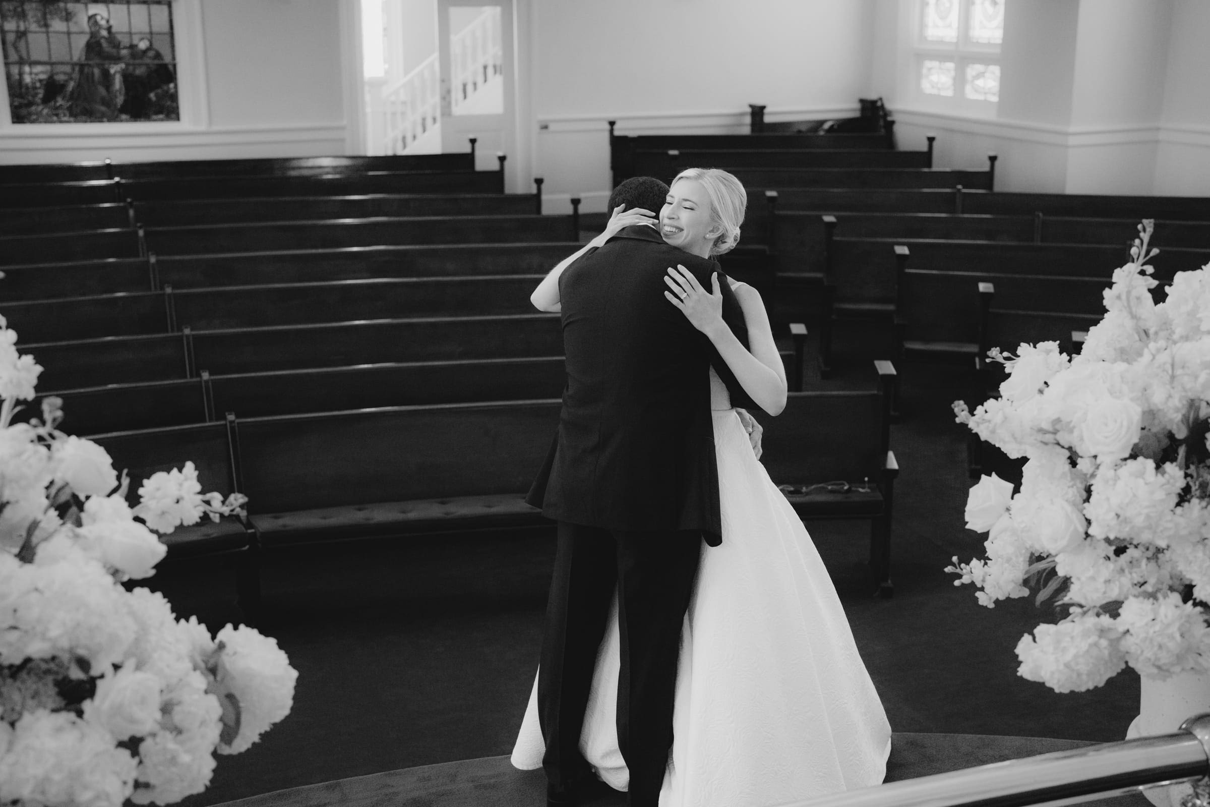 dunn wedding photography - reaves & zach A black-and-white photo by Dunn wedding photography captures a bride and groom embracing in a church. The bride wears a white dress, the groom a dark suit. Surrounded by floral arrangements, the scene is beautifully framed with the church pews visible in the background.