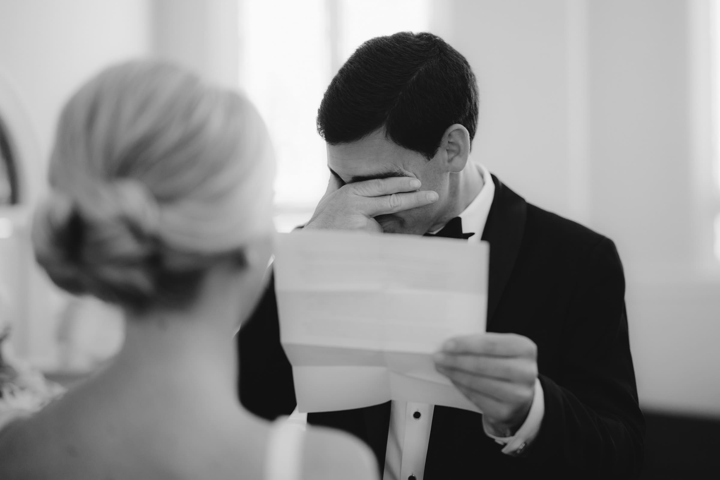 dunn wedding photography - reaves & zach A groom in a tuxedo wipes his eyes while reading a letter, visibly emotional, as captured by Dunn wedding photography. A bride with her hair in an elegant bun stands nearby, seen from behind. The black-and-white scene beautifully captures this heartfelt moment.