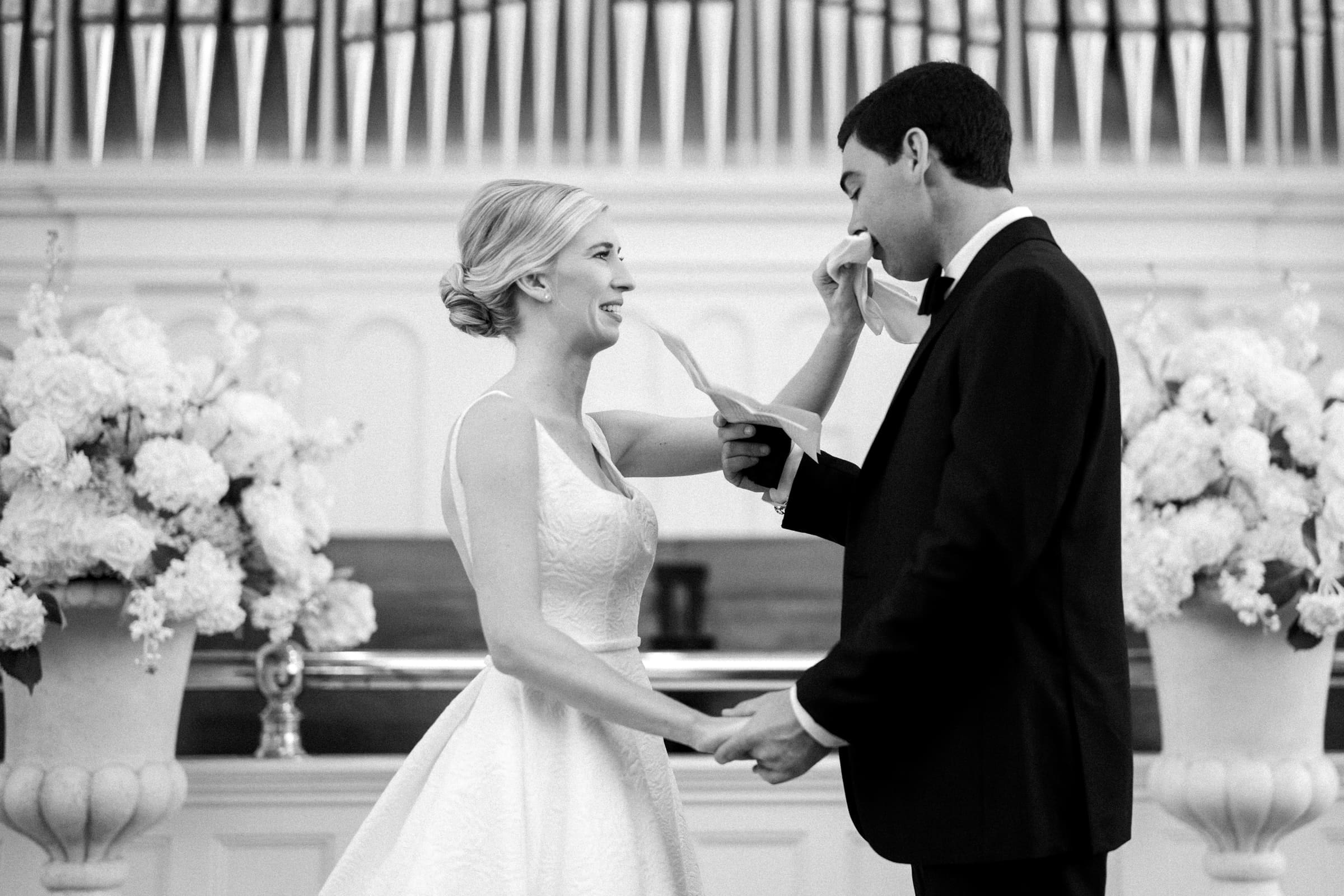 dunn wedding photography - reaves & zach The bride and groom smile at each other, holding hands in front of a decorated altar. The groom wipes his eyes with a handkerchief as the bride speaks. Captured by Dunn Wedding Photography, large flower arrangements and an organ create the perfect backdrop.