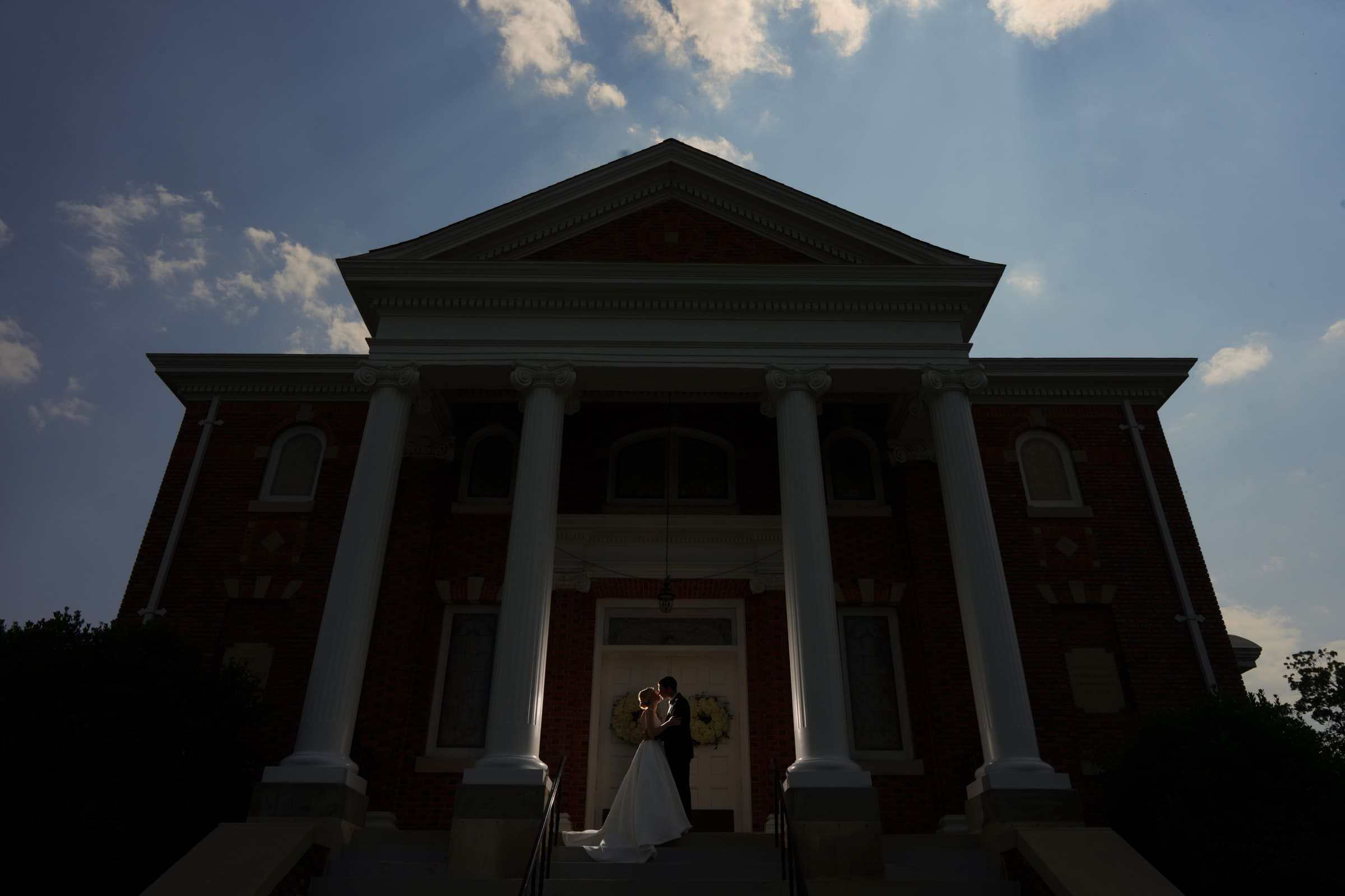 dunn wedding photography - reaves & zach A couple stands in an embrace at the entrance of a large, sunlit brick church with tall white columns, perfectly captured by Dunn wedding photography. Silhouetted against the bright sky and surrounded by soft, ambient light, their love is beautifully highlighted.