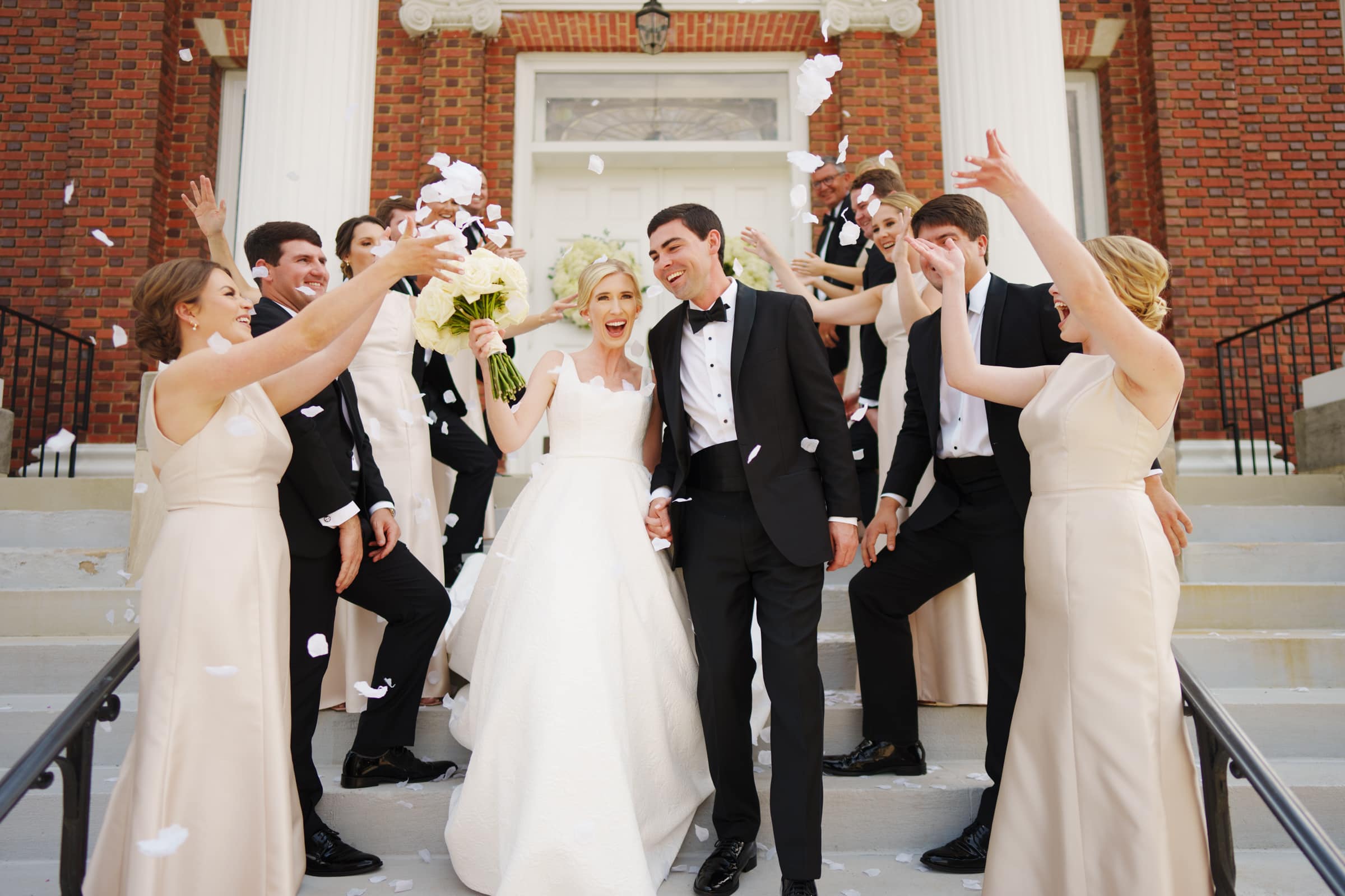 dunn wedding photography - reaves & zach A bride and groom in formal attire smile joyfully on steps with their wedding party, captured perfectly by Dunn Wedding Photography. Bridesmaids in beige dresses and groomsmen in black tuxedos celebrate, throwing white flower petals into the air, with a brick building standing majestically behind them.