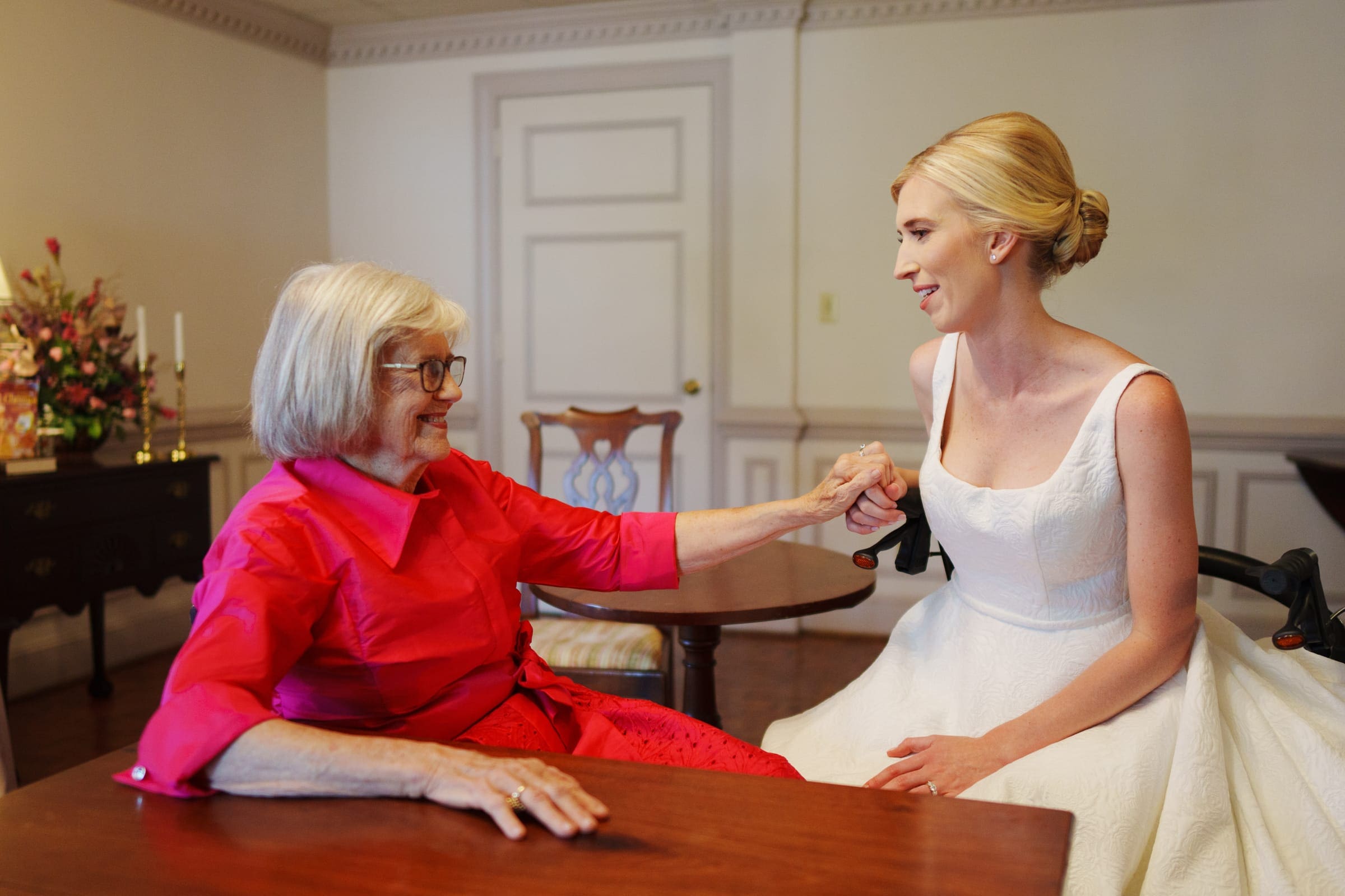 dunn wedding photography - reaves & zach A bride in a white dress holds hands with an older woman in a red outfit, both smiling at each other in a warmly lit room. Wooden furniture and a floral arrangement enhance the ambiance captured by Dunn wedding photography.