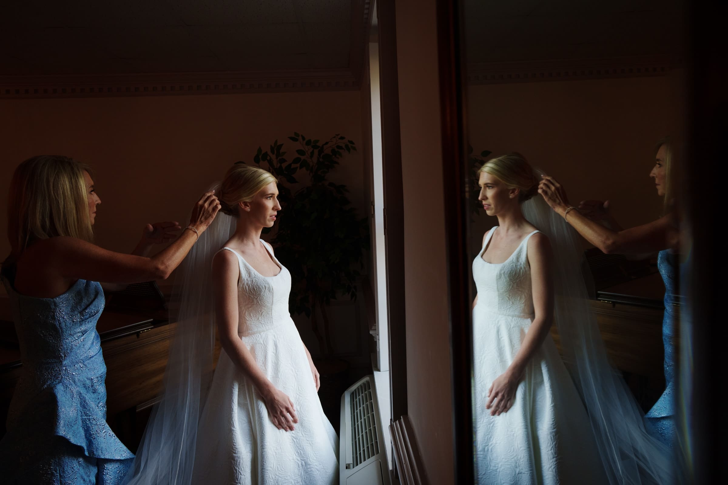 dunn wedding photography - reaves & zach A bride in a white dress and veil stands by a window, her reflection captured perfectly by Dunn wedding photography. Another woman, dressed in blue, gently adjusts the bride's veil. The room is dimly lit, with lush plants adding to the serene ambiance.
