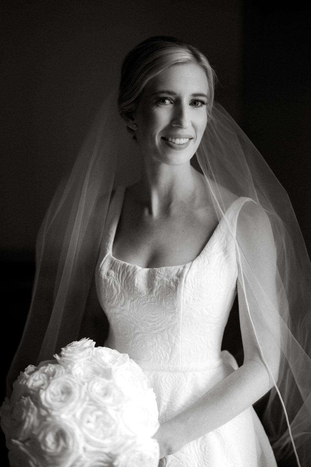 dunn wedding photography - reaves & zach A bride in an elegant, textured white gown smiles softly, holding a bouquet of white flowers. Her veil gently drapes over her shoulders. Captured by Dunn wedding photography, the black and white image highlights the delicate details of her attire and bouquet.