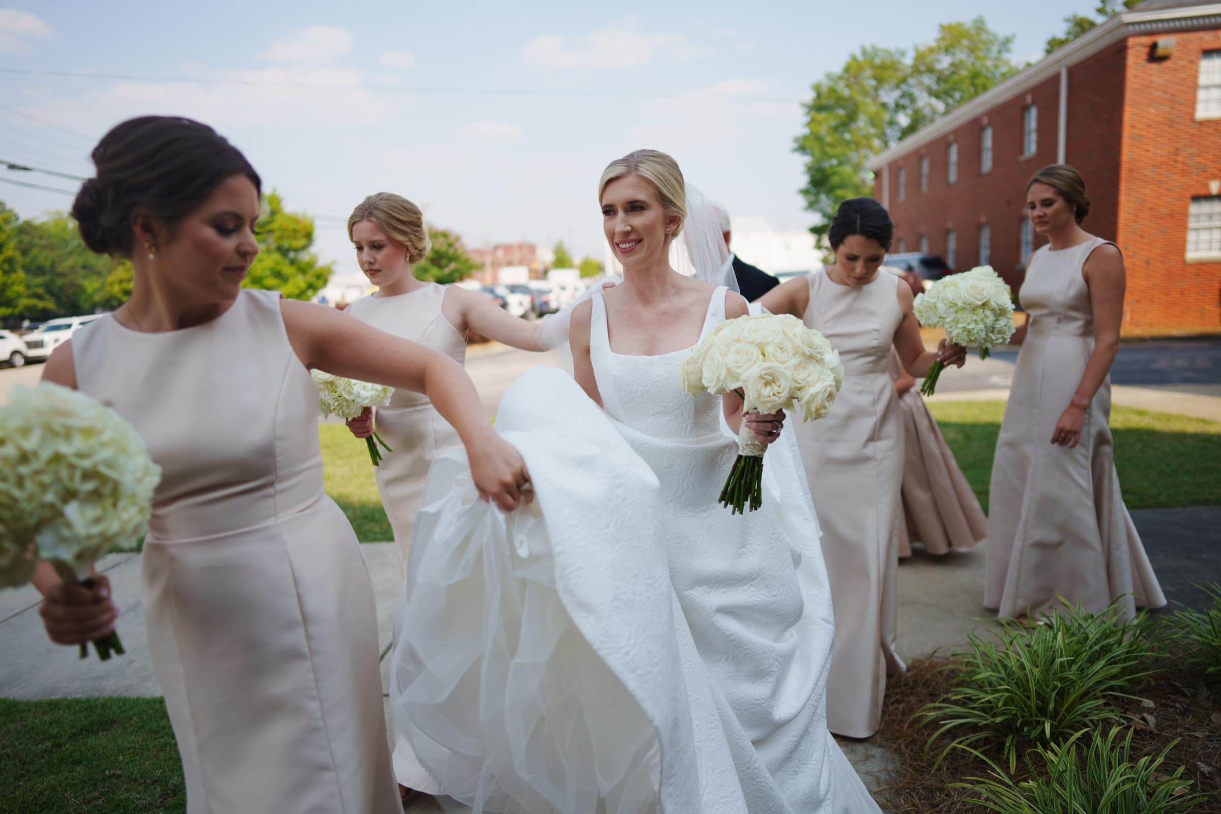 dunn wedding photography - reaves & zach A bride in a white gown, holding a bouquet of white roses, walks with her bridesmaids in matching beige dresses. Captured by Dunn Wedding Photography, they are outside on a sunny day, with one bridesmaid helping carry the bridal train. A red brick building is in the background.