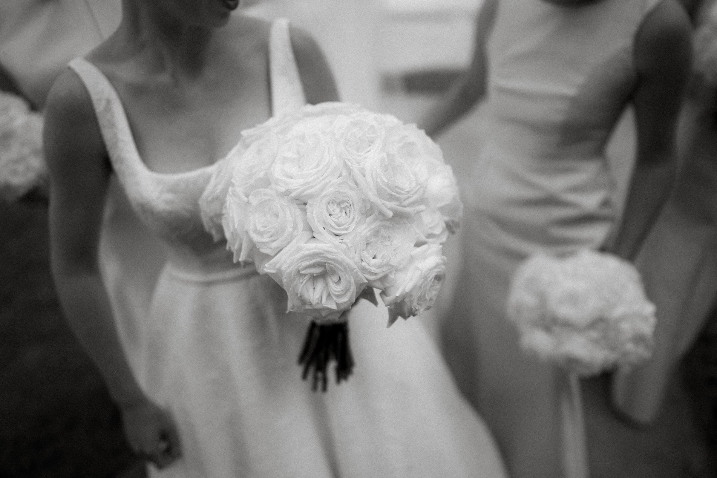 dunn wedding photography - reaves & zach A black and white image captures two individuals in elegant, sleeveless dresses, each cradling a bouquet of white roses. The focus on the foreground bouquet exudes timeless beauty—a hallmark of Dunn wedding photography—while the softly blurred background adds a dreamlike quality.