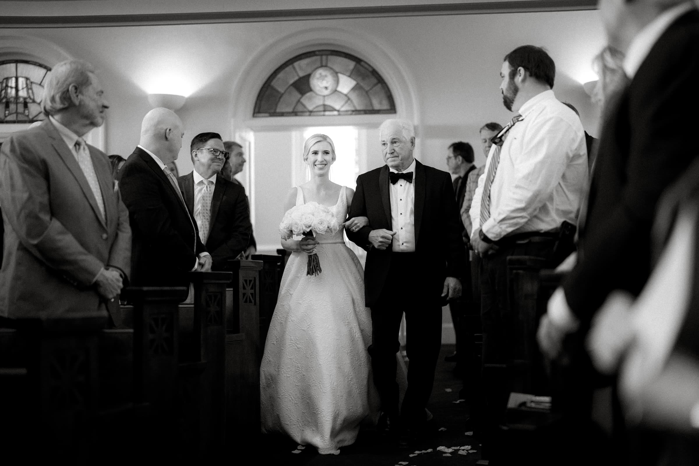 dunn wedding photography - reaves & zach A bride, clutching a bouquet, walks down the aisle with an older man in a tuxedo, captured perfectly by Dunn wedding photography. Guests in formal attire look on as they pass through the church's arched-window backdrop.