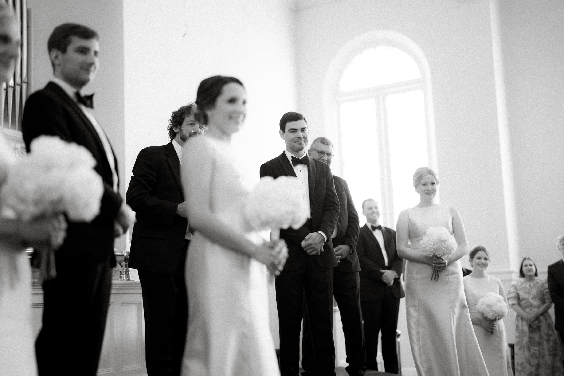 dunn wedding photography - reaves & zach A black and white photo of a formal indoor gathering, possibly a wedding captured by Dunn wedding photography. Several people in suits and dresses stand near a large arched window, bouquets in hand, radiating an atmosphere of anticipation and joy.