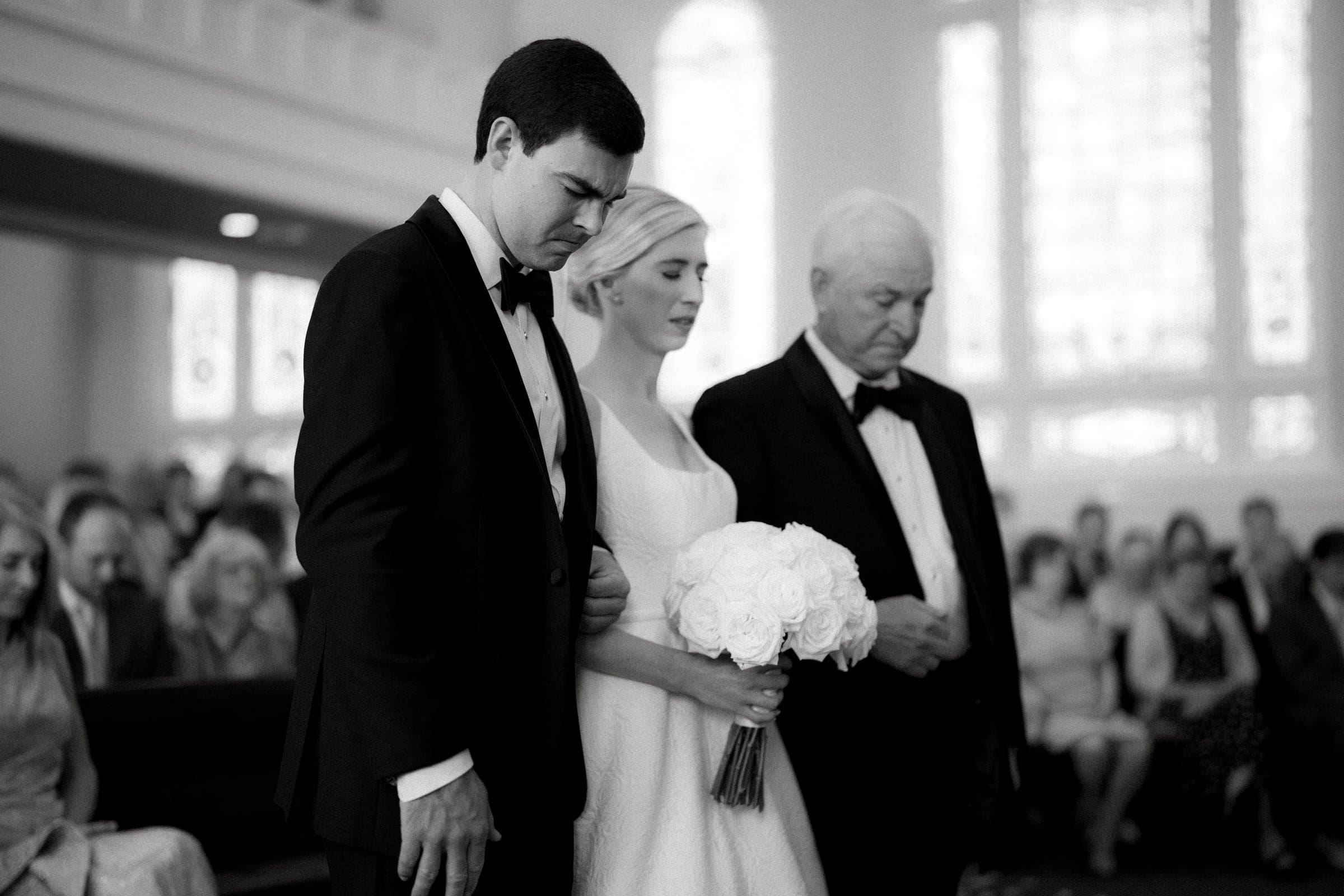 dunn wedding photography - reaves & zach A black and white photo captures a solemn moment at a Dunn wedding: the bride and groom, standing with an older man in formal attire, likely during the ceremony. The bride clutches a bouquet of white flowers, surrounded by seated guests in a church.
