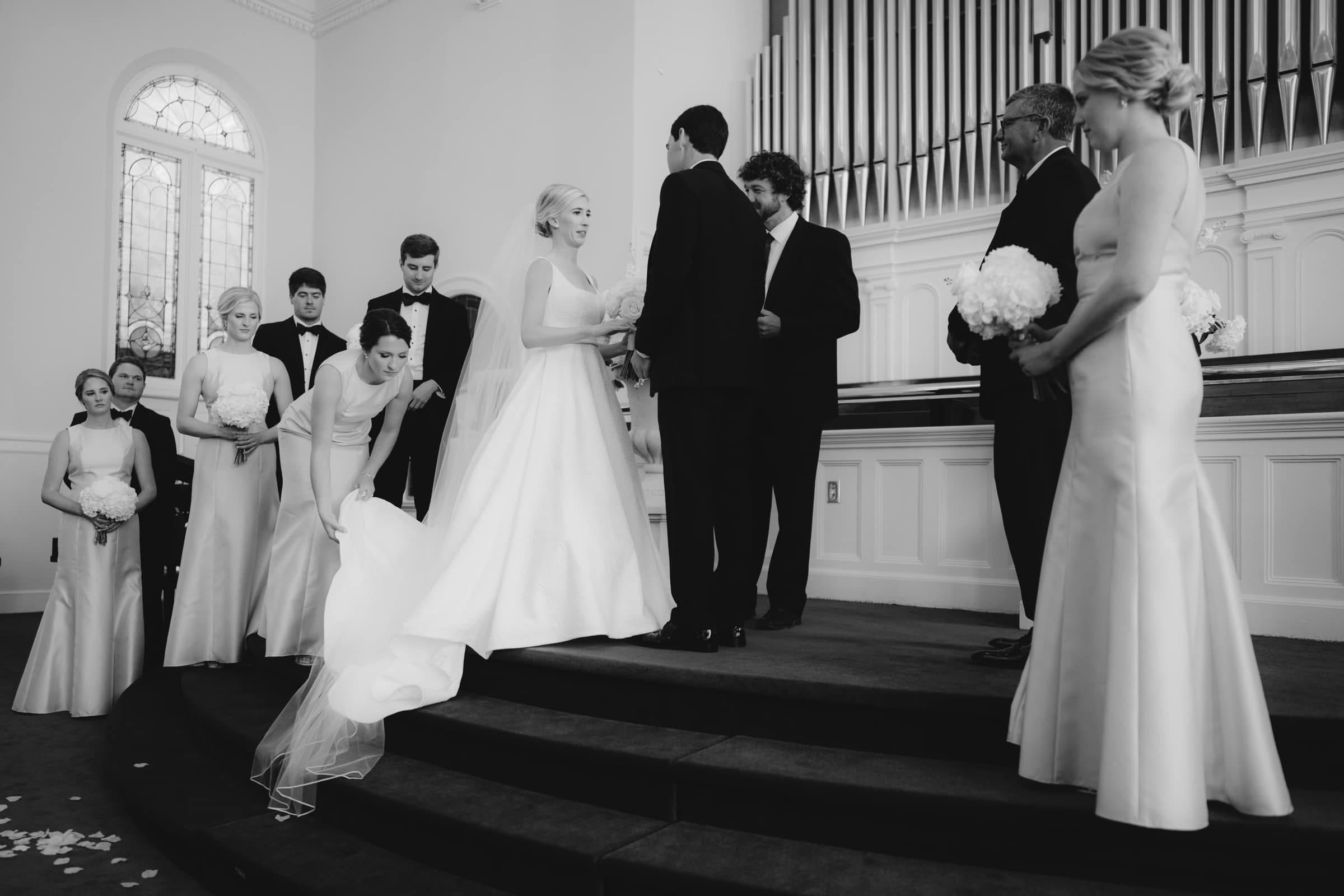 dunn wedding photography - reaves & zach A black and white photo captures the Dunn wedding ceremony. The bride and groom stand on a platform, exchanging rings, as bridesmaids and groomsmen surround them. One bridesmaid adjusts the bride's dress train. The venue boasts a large pipe organ and stained glass windows, enhancing the timeless moment.