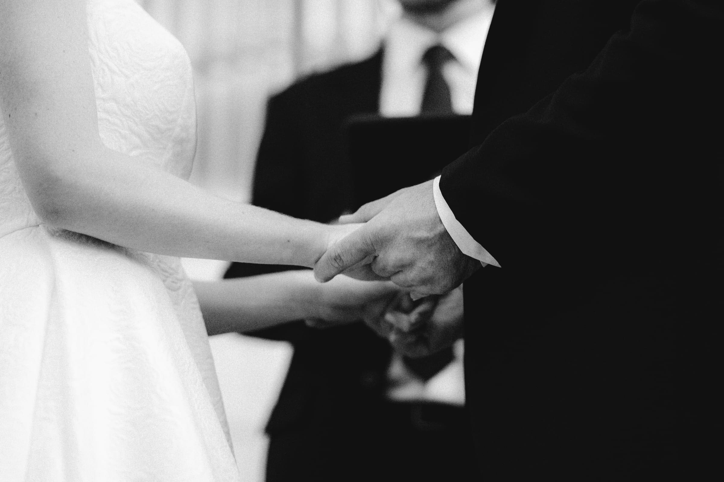 dunn wedding photography - reaves & zach A timeless black and white capture by Dunn wedding photography shows a couple holding hands during their ceremony. The bride wears a textured white dress while the groom dons a suit. In the background, someone holds a book, likely the officiant, adding depth to this cherished moment.