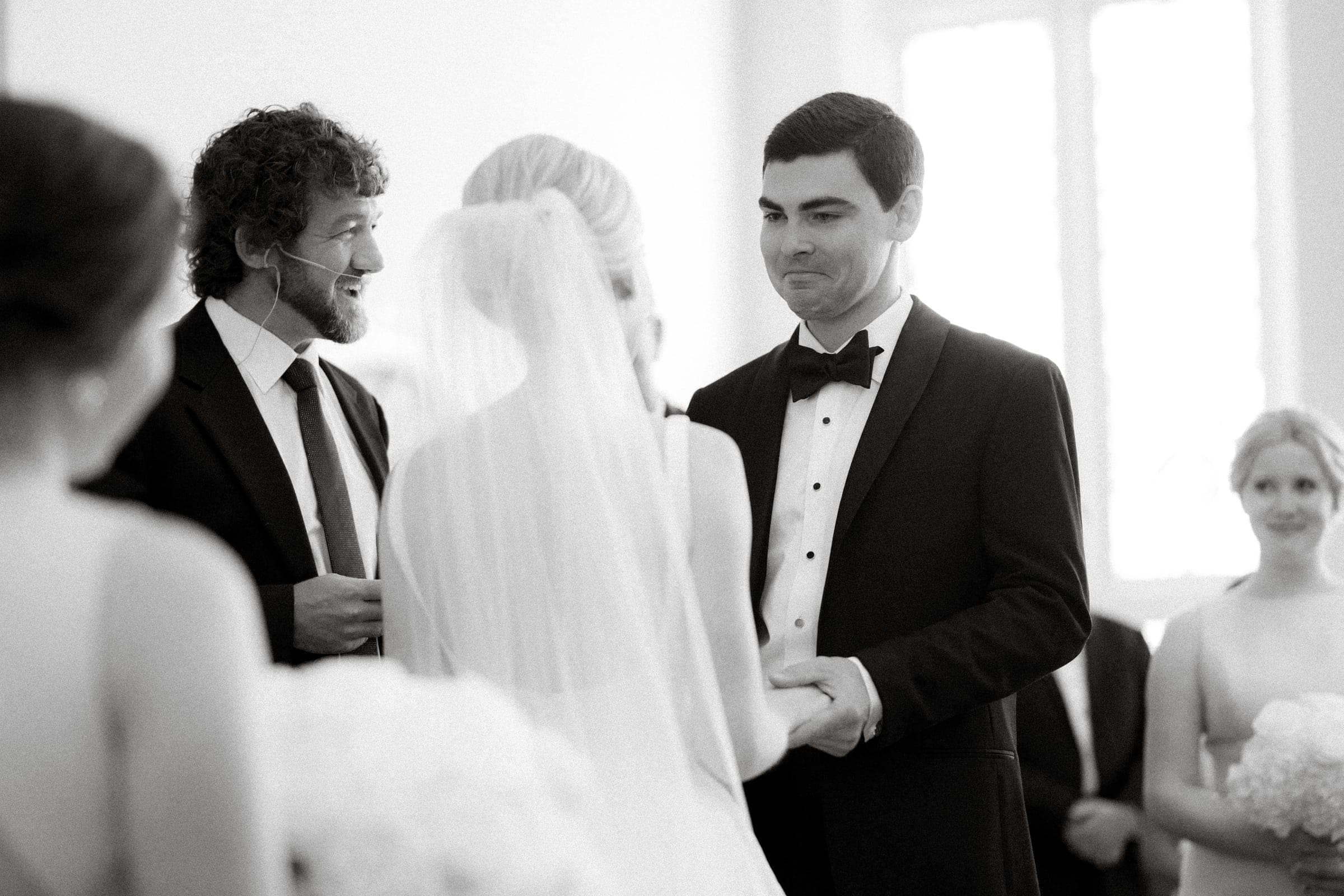 dunn wedding photography - reaves & zach A black and white photo by Dunn wedding photography captures a bride and groom standing hand in hand, facing each other lovingly. The officiant smiles warmly, while another person watches from the background, adding to the timeless charm of the moment.