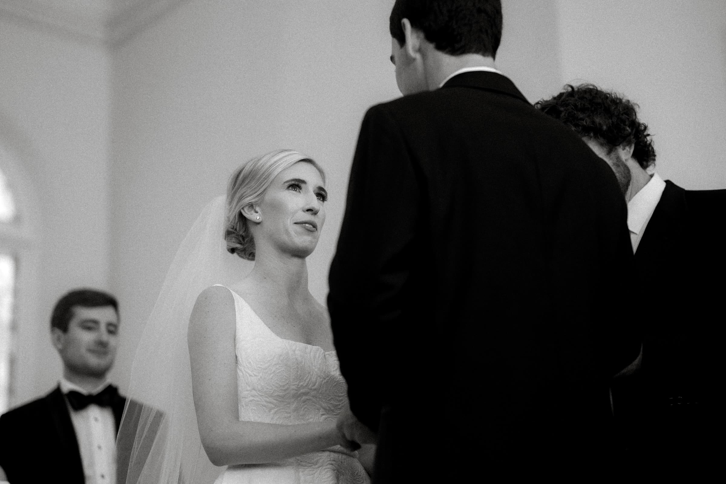 dunn wedding photography - reaves & zach A black and white photo captures a bride and groom facing each other during a Dunn wedding ceremony. The bride's veil gracefully complements her white dress, while the groom stands in a dark suit. In the background, two men in suits are subtly visible, adding depth to this timeless moment.