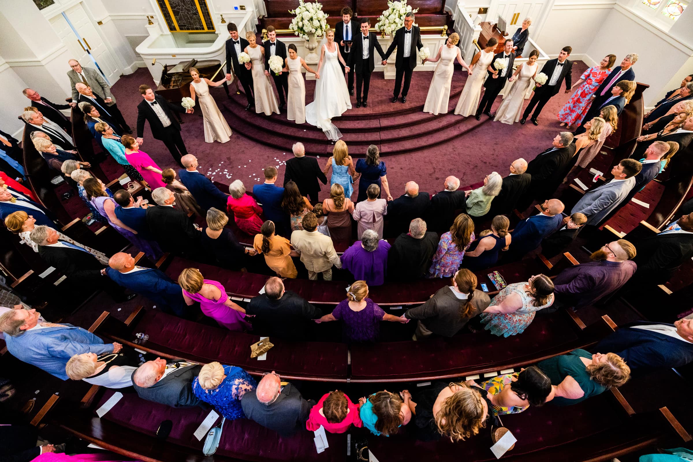 dunn wedding photography - reaves & zach A birds-eye view of the Dunn wedding ceremony inside a church captures the bride and groom at the altar, surrounded by a semicircle of bridesmaids and groomsmen. Guests fill the pews, watching the couple as the church is adorned with flowers.