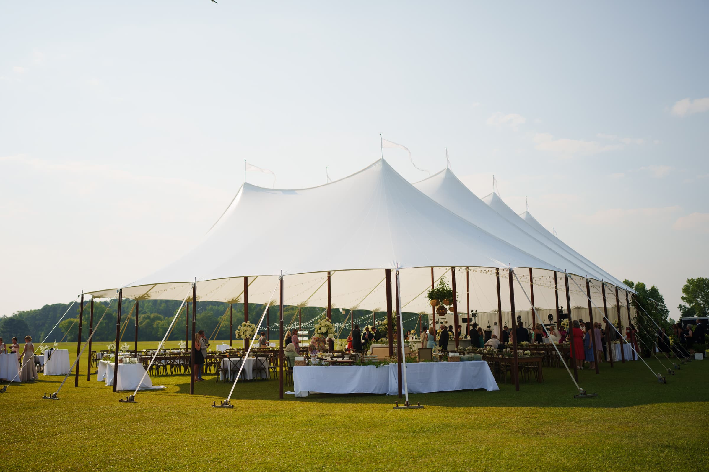 dunn wedding photography - reaves & zach A large white event tent set up on a grassy field under a clear blue sky, with several tables and people gathered inside, beautifully captured by Dunn Wedding Photography, suggesting a celebration or gathering.
