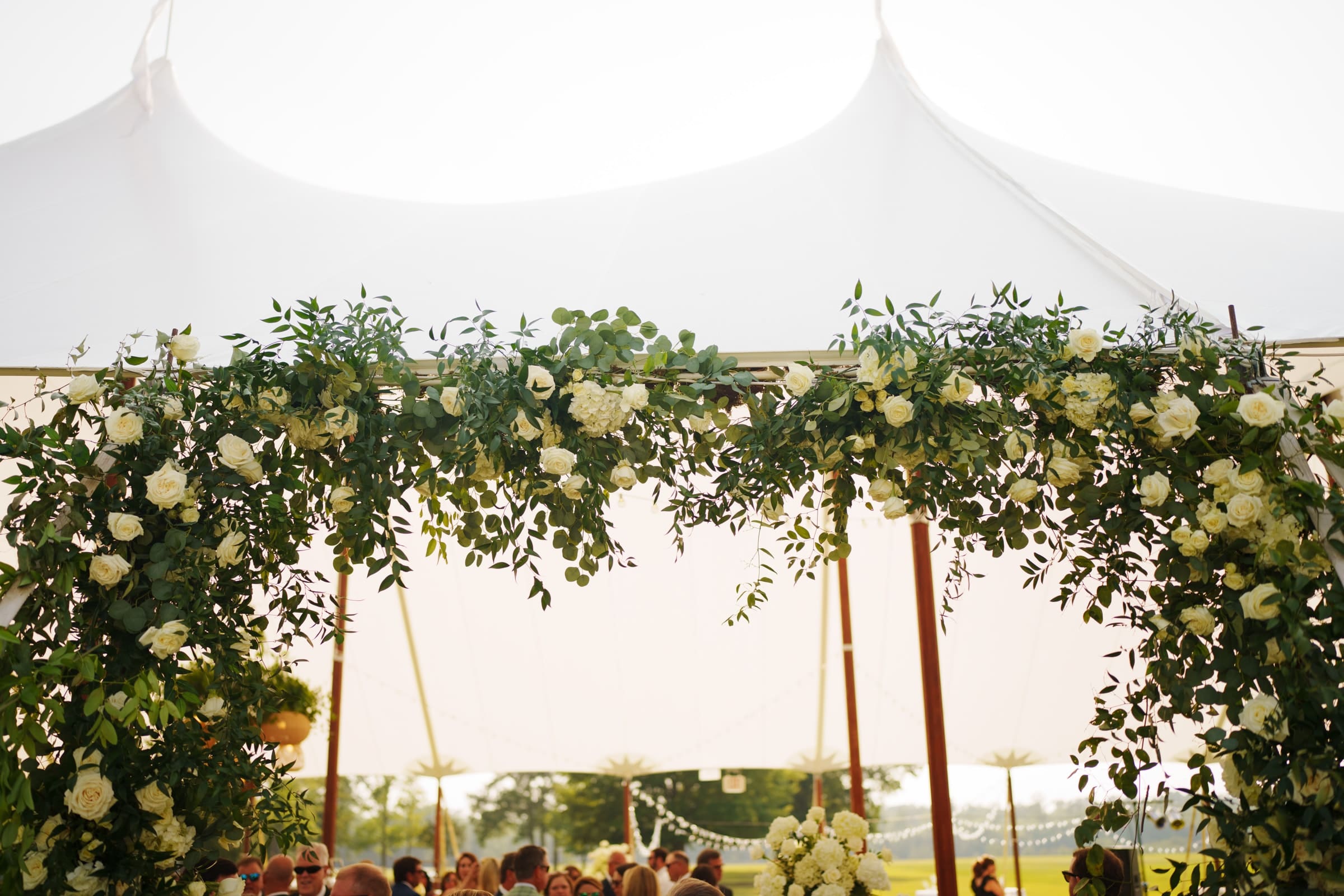 dunn wedding photography - reaves & zach A white outdoor tent adorned with greenery and white flowers forms a picturesque arch over the gathering, skillfully captured by Dunn wedding photography. The foreground brims with lush foliage against a backdrop of bright, sunny skies.