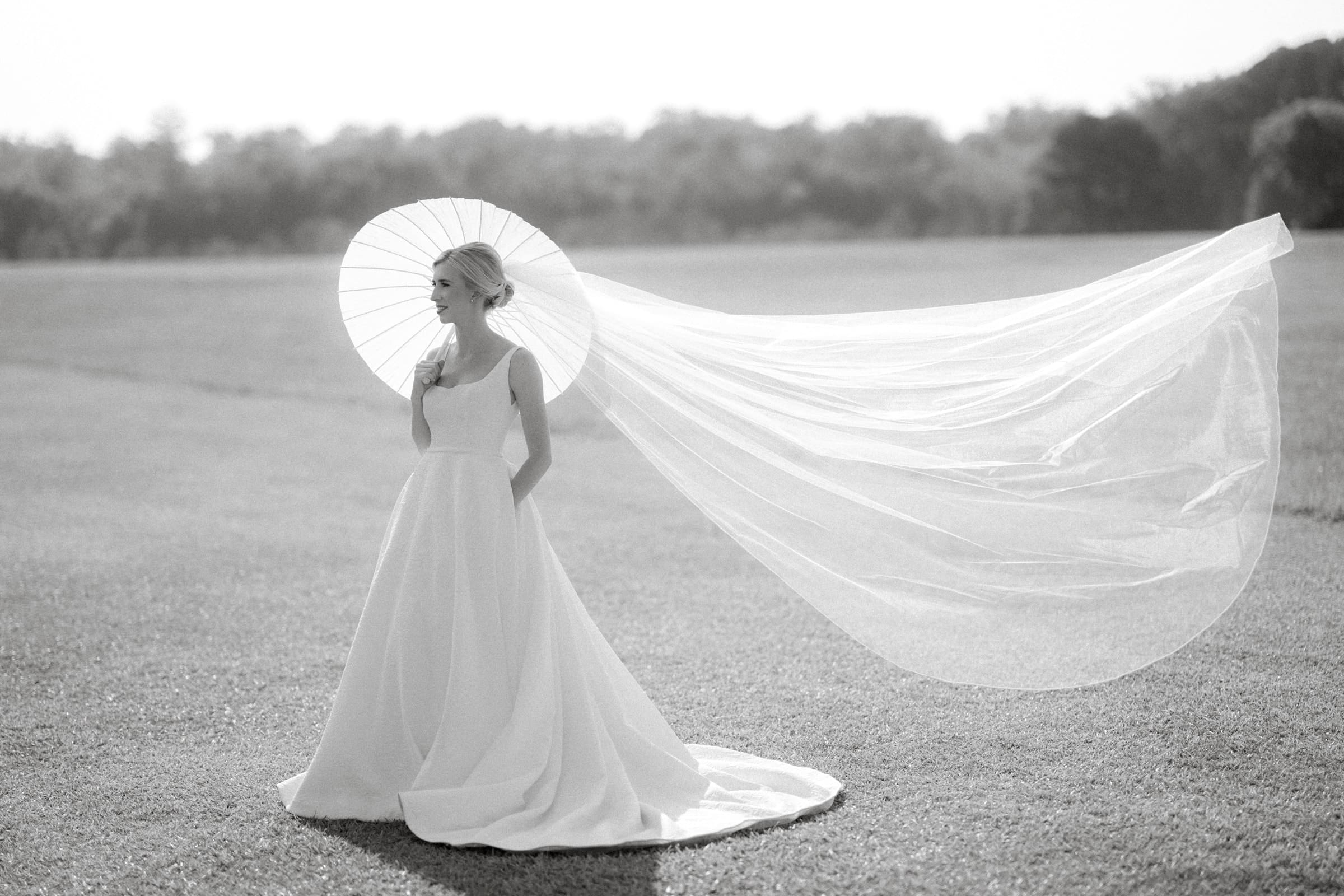 dunn wedding photography - reaves & zach A bride stands gracefully on a grassy field, wearing a flowing white gown and a long, flowing veil that billows in the breeze. Captured by Dunn Wedding Photography, she holds a delicate parasol over her shoulder against the soft-focus backdrop of trees.