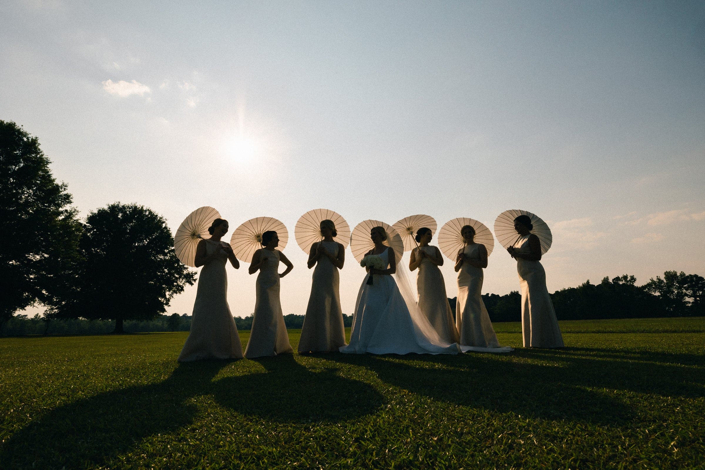 dunn wedding photography - reaves & zach At the Dunn wedding, a bride and her bridesmaids gracefully hold parasols in a grassy field under a clear sky. The sun casts long shadows while a tree stands peacefully in the background, beautifully captured through exquisite photography.
