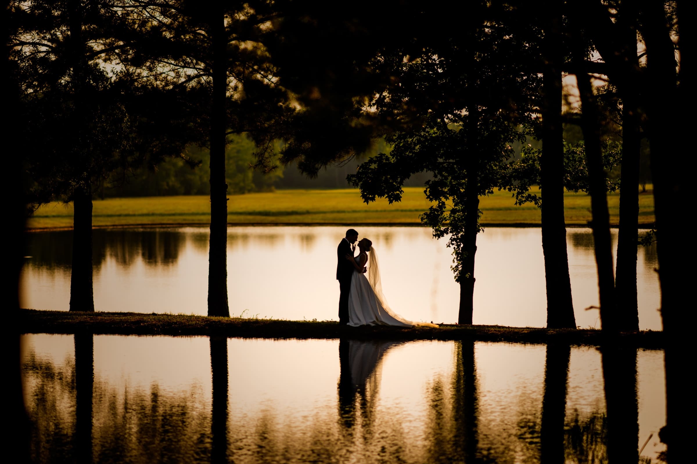 A couple embraces on a narrow strip of land surrounded by a peaceful lake, silhouetted against a warm sunset. Tall trees frame the scene, their reflections crafting a serene and romantic atmosphere—an idyllic moment captured by Dunn Wedding Photography.