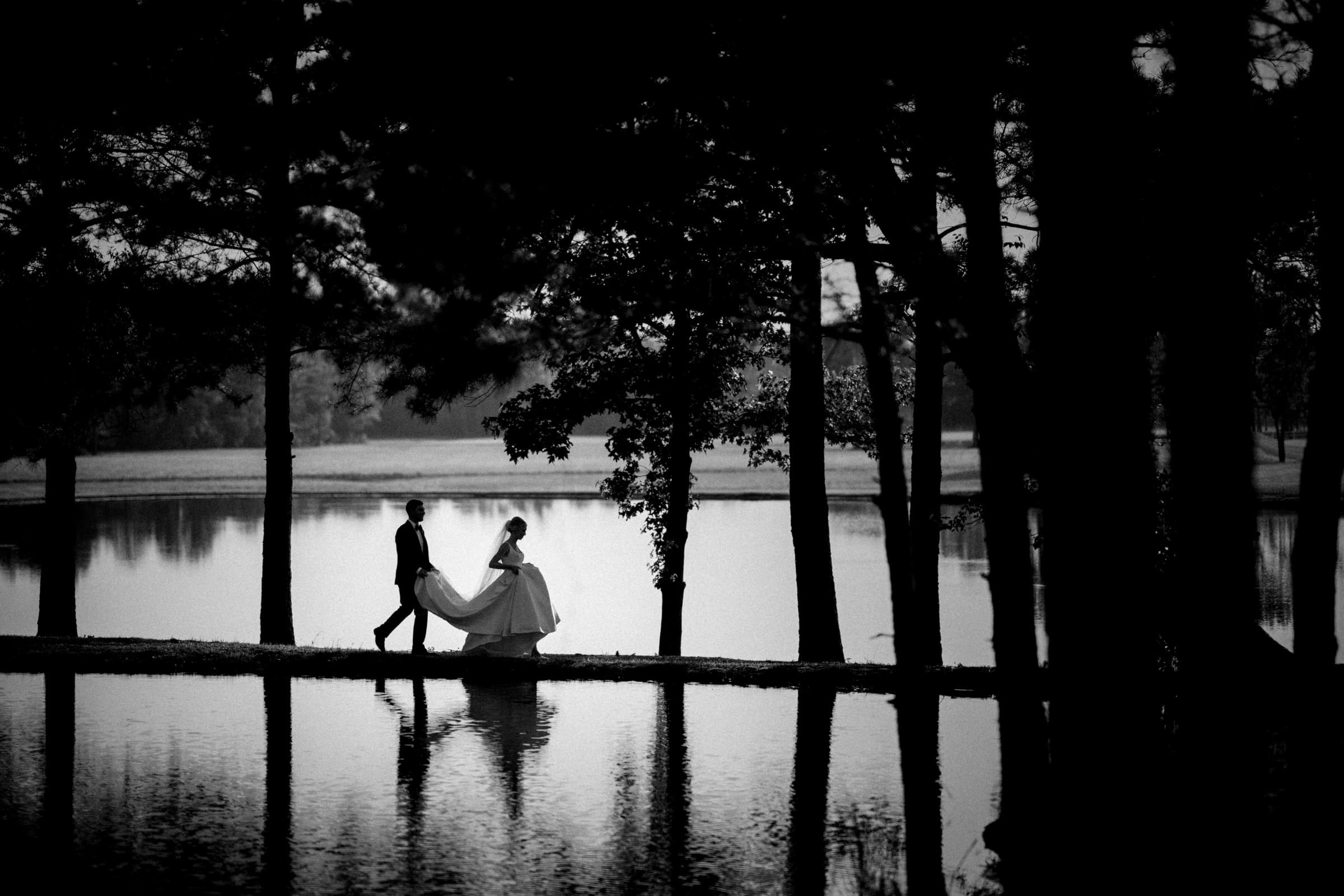 dunn wedding photography - reaves & zach A couple in wedding attire walks hand in hand beside a scenic lake, framed by tall trees. The bride's gown flows elegantly behind her, and the water reflects their silhouettes, crafting a serene and romantic atmosphere captured beautifully by Dunn Wedding Photography.