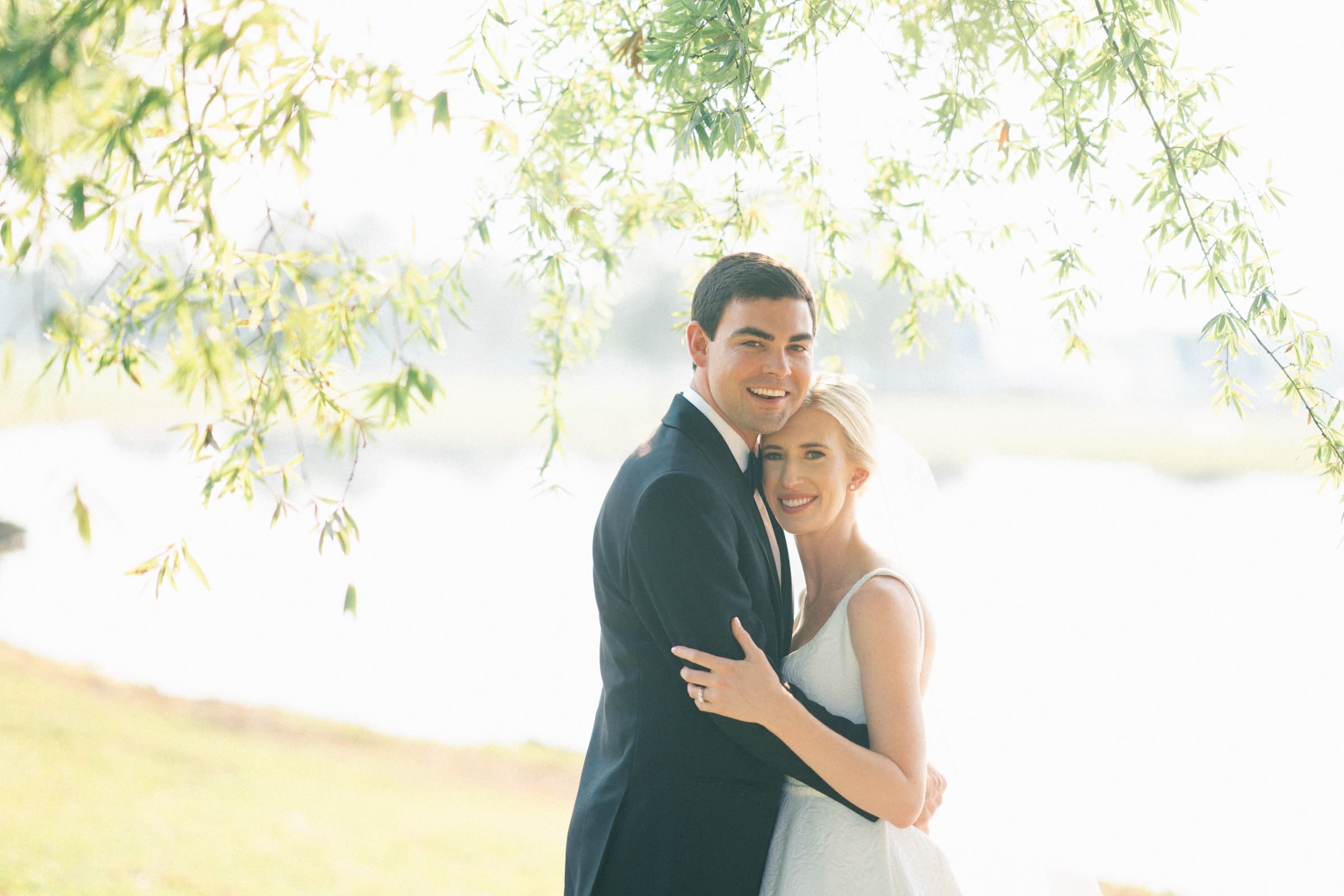 dunn wedding photography - reaves & zach A bride and groom smile warmly under a tree with green leaves in this Dunn wedding photography masterpiece. The groom wears a black suit, and the bride is in a white dress, embracing each other with a blurred, sunlit background.