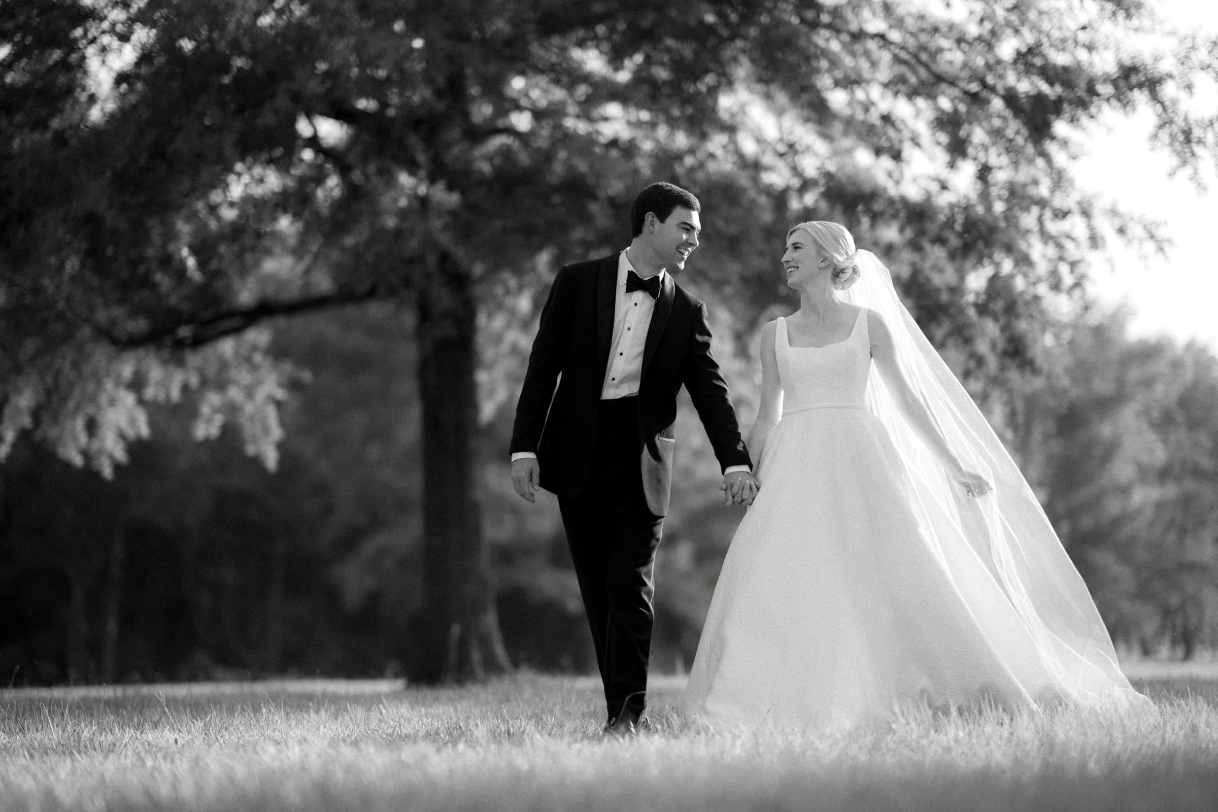 dunn wedding photography - reaves & zach A groom in a tuxedo and a bride in a wedding dress walk hand in hand, perfectly framed by the artistry of Dunn wedding photography. They are in a grassy area with large trees in the background, smiling at each other. The scene is captured beautifully in black and white.
