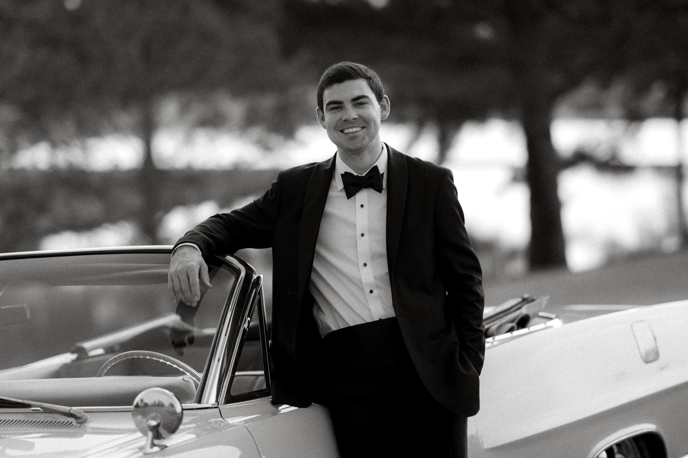 dunn wedding photography - reaves & zach A man in a tuxedo smiles while leaning on a vintage convertible car, captured beautifully by Dunn Wedding Photography. The background features blurred trees and what appears to be a body of water, all elegantly presented in black and white.