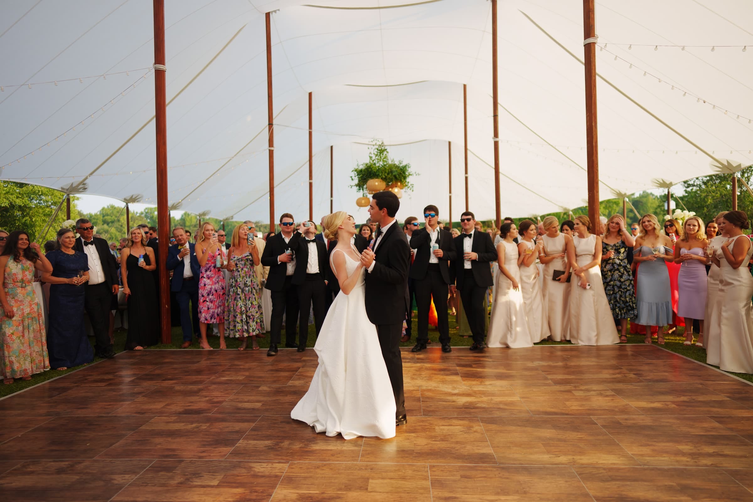 dunn wedding photography - reaves & zach A couple is dancing under a large white tent, captured perfectly by Dunn wedding photography, as elegantly dressed guests clap and watch. The wooden dance floor gleams with greenery at the edges, enhancing the festive and celebratory atmosphere.