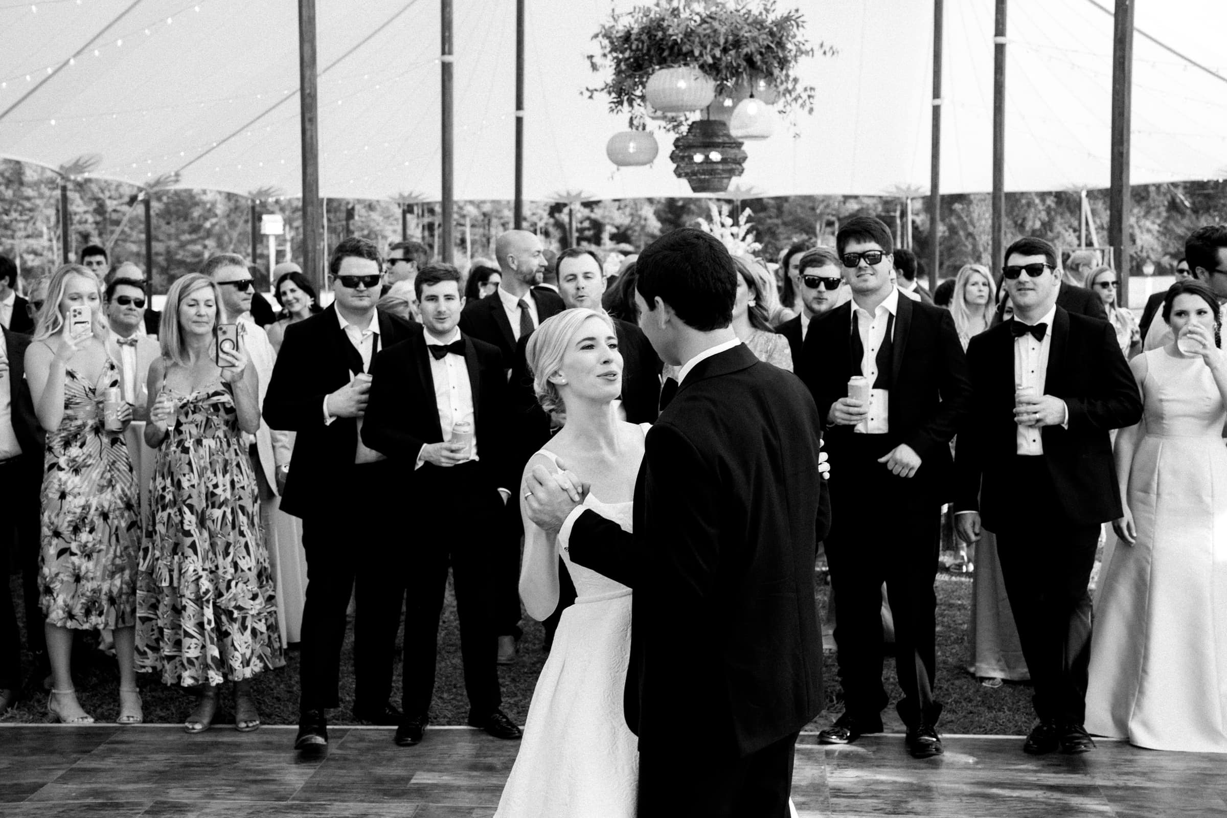dunn wedding photography - reaves & zach A black and white photo captures a couple dancing at a wedding reception. Guests, some holding drinks, watch and smile in the background. The event unfolds in a tent adorned with string lights and hanging decorations, showcasing Dunn wedding photography at its finest.