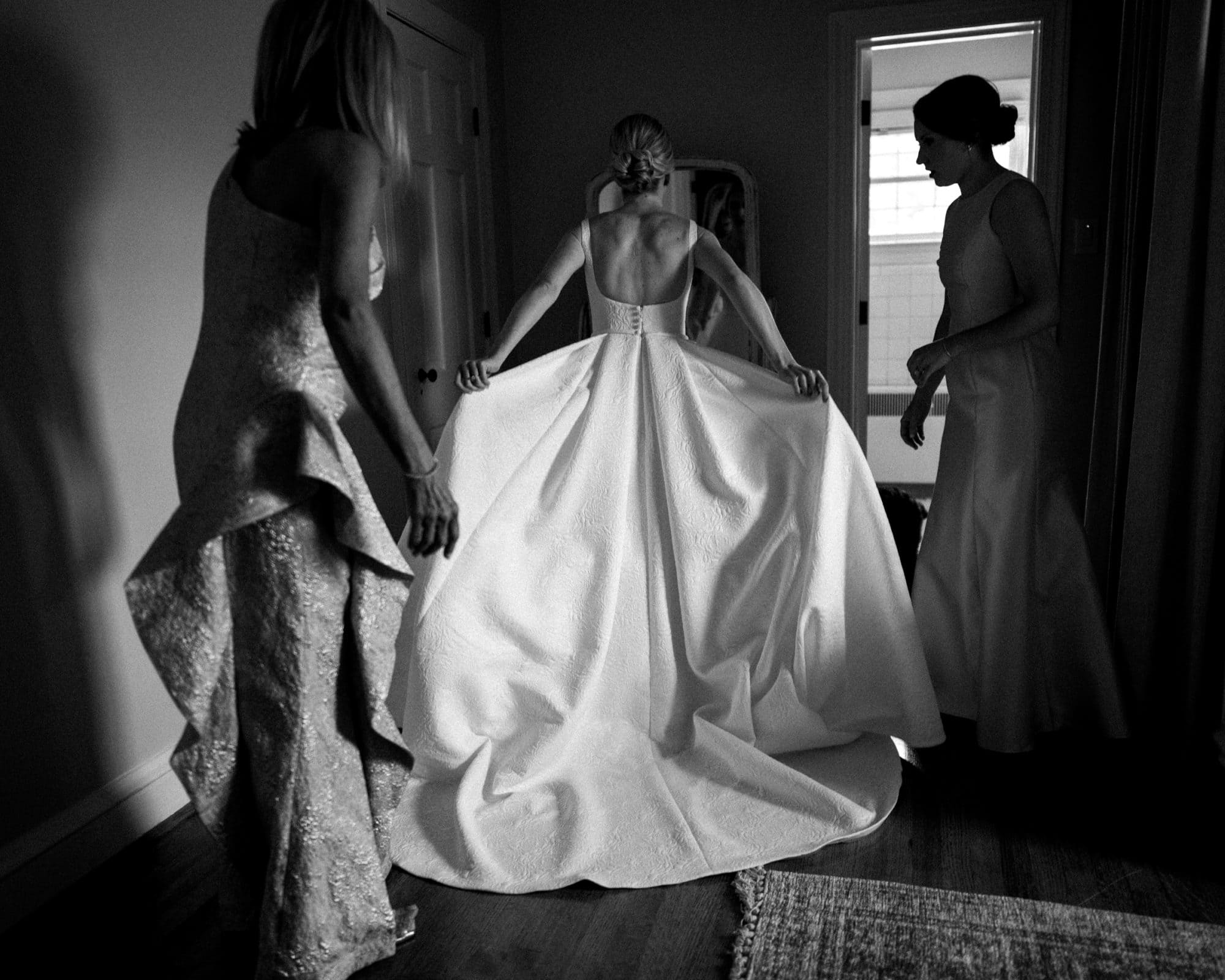 dunn wedding photography - reaves & zach In a softly lit room, a bride in her wedding gown adjusts her dress with the help of two women. This intimate moment before the ceremony is beautifully captured, showcasing the artistry of Dunn wedding photography.