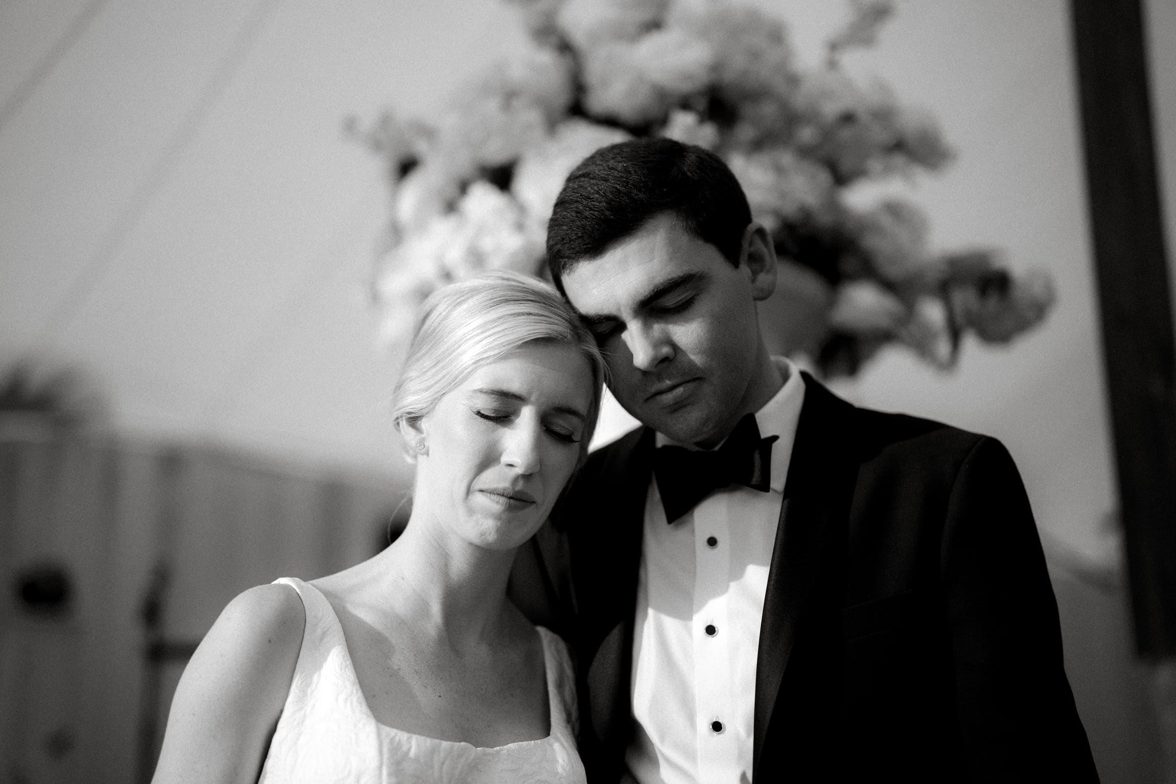 dunn wedding photography - reaves & zach A black and white photo by Dunn Wedding Photography captures a bride and groom standing closely with eyes closed, dressed in wedding attire. The bride wears a sleeveless dress, while the groom is in a tuxedo. A blurred floral arrangement is visible in the background.