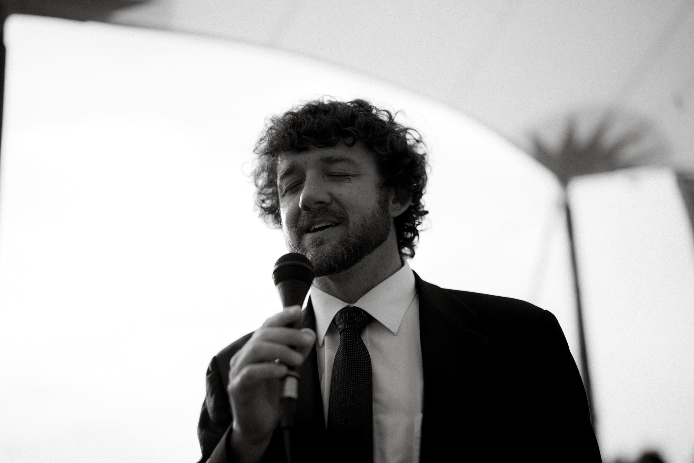 dunn wedding photography - reaves & zach A man with curly hair and a beard, dressed in a suit and tie, holds a microphone with eyes closed, exuding emotion as if lost in song or speech. The softly blurred background hints at a tented setting, capturing the timeless elegance typical of Dunn wedding photography. Black and white photo.