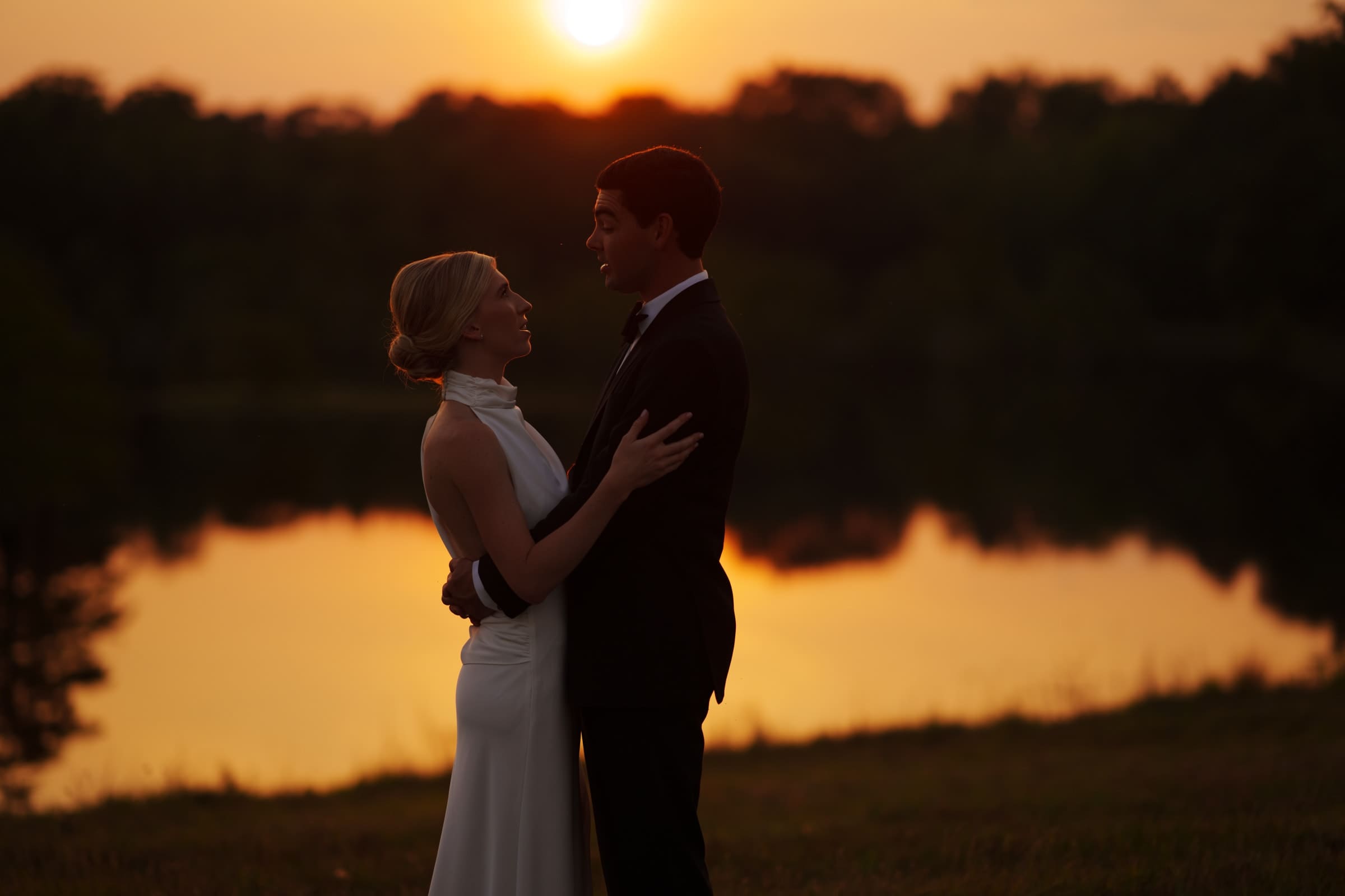 dunn wedding photography - reaves & zach A couple stands in an embrace by a calm lake at sunset, the orange and yellow hues illuminating the sky and reflecting on the water. Trees line the background, enhancing the serene and romantic atmosphere captured perfectly by Dunn Wedding Photography.