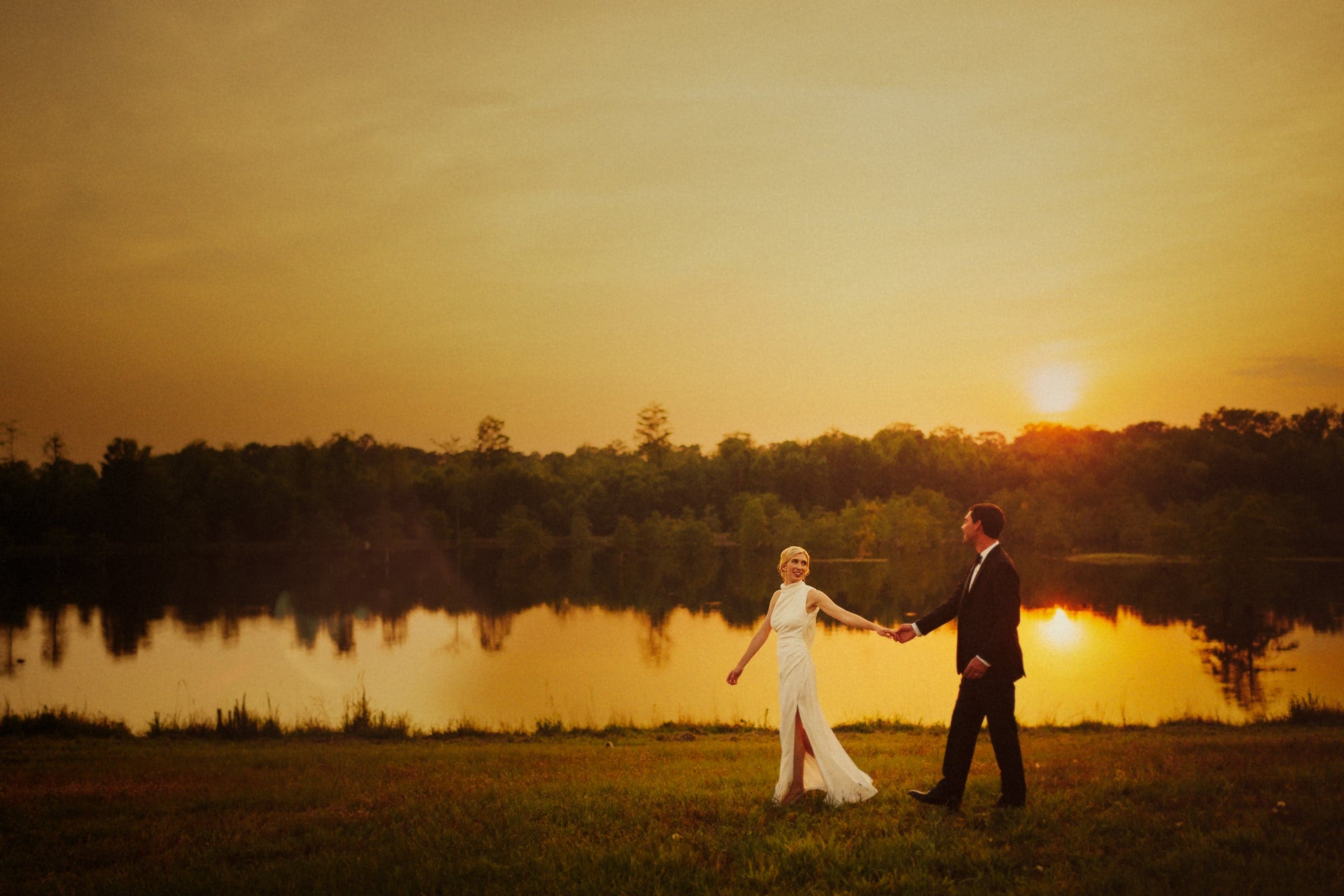 dunn wedding photography - reaves & zach A couple in formal attire walks hand in hand near a lake at sunset, captured perfectly by Dunn wedding photography. The sky glows with warm hues, reflecting in the water. The woman wears a white dress and the man a dark suit, creating a serene and romantic atmosphere.