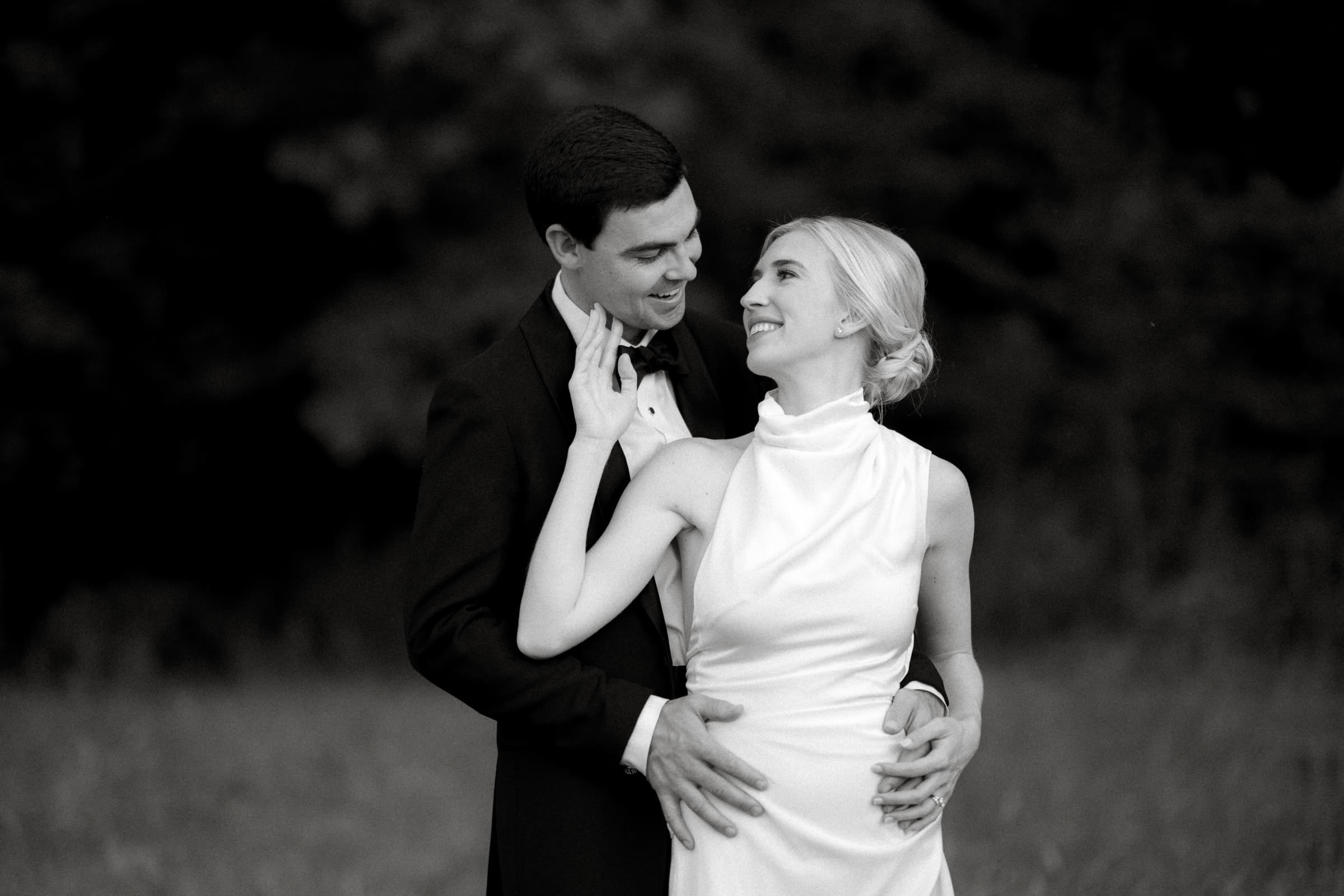dunn wedding photography - reaves & zach A black and white photo captures a couple in formal attire, the man in a suit and the woman in a sleeveless dress, smiling at each other amidst an outdoor setting with trees. This timeless moment exemplifies Dunn wedding photography’s signature style.