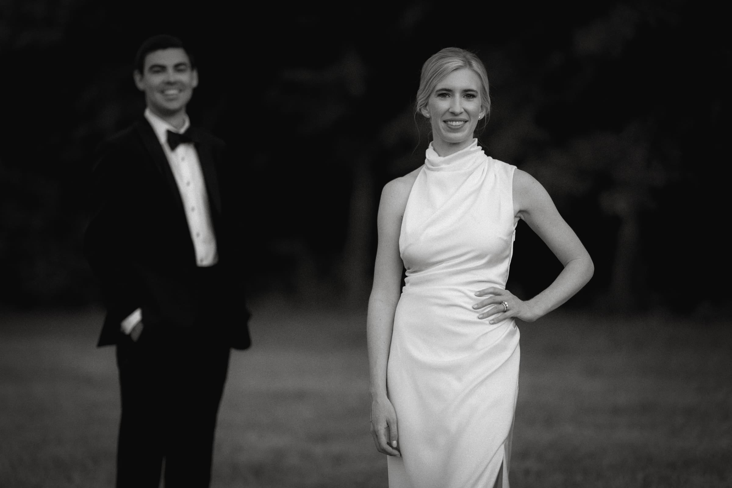 dunn wedding photography - reaves & zach A grayscale photo captures a woman in a white gown, smiling with her hand on her hip, embodying timeless elegance typical of Dunn wedding photography. In the background, a man in a tuxedo stands with his hands in his pockets amidst an outdoor setting of grass and trees.