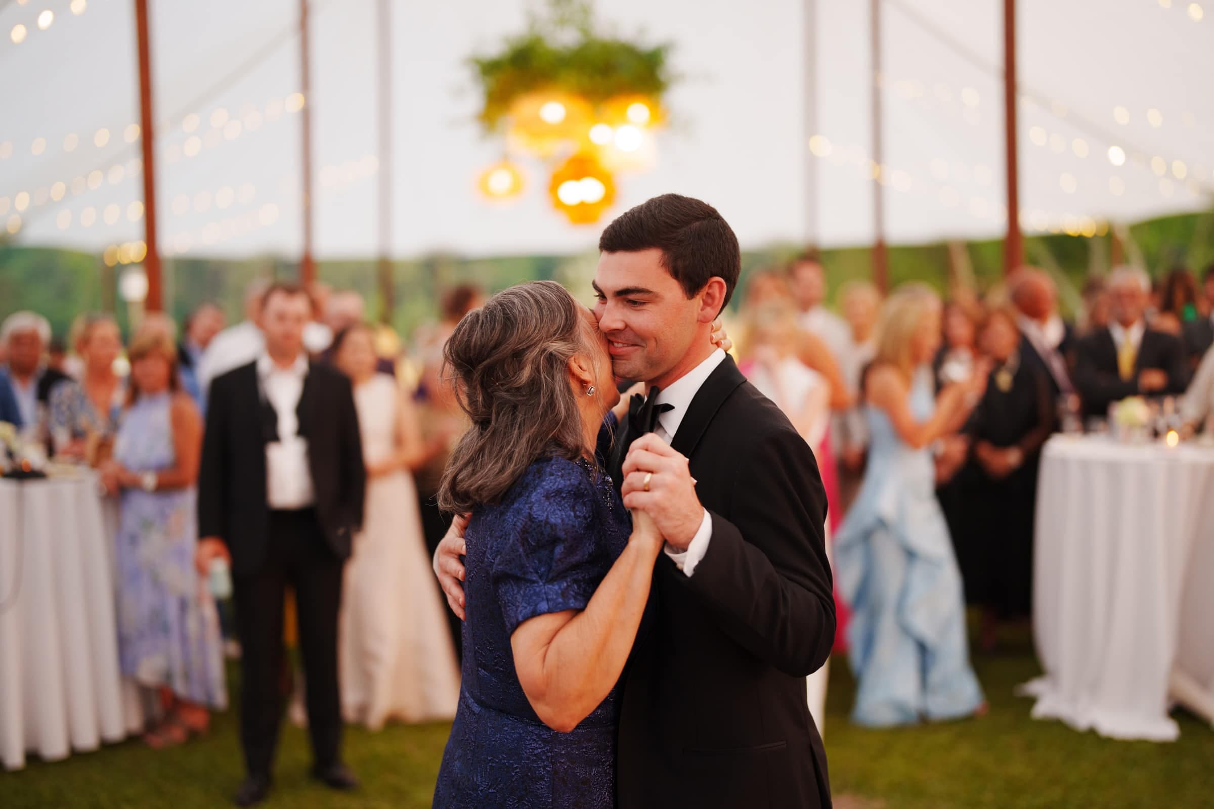 dunn wedding photography - reaves & zach A man in a suit and a woman in a blue dress dance closely under a tent adorned with string lights, captured beautifully by Dunn wedding photography. A crowd in formal attire watches them, enveloped in the festive and joyful atmosphere.