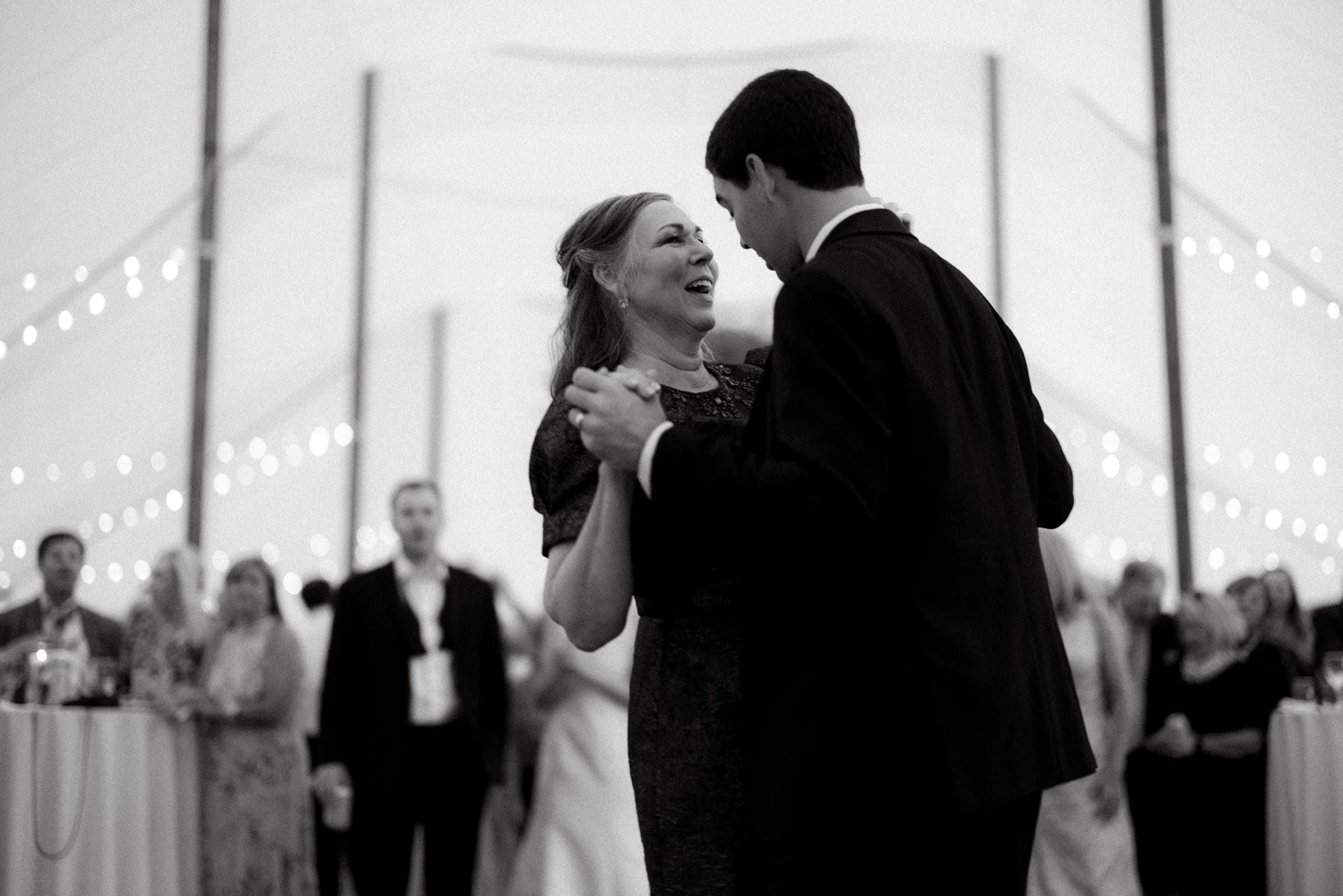 dunn wedding photography - reaves & zach A couple dances closely under a tent, surrounded by guests. The woman is smiling in a dark dress, while the man wears a suit. Soft lights strung above create a warm, nostalgic atmosphere captured brilliantly in black and white by Dunn Wedding Photography.