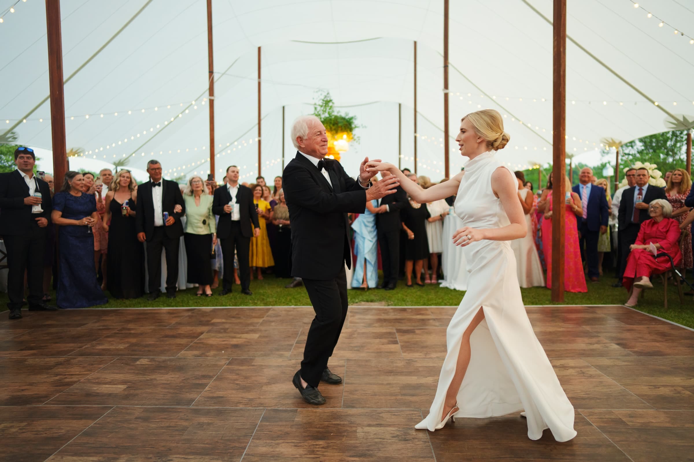 dunn wedding photography - reaves & zach A couple elegantly dances on a wooden floor under a large, white marquee tent adorned with string lights, captured in stunning Dunn wedding photography. The woman wears a white dress with a slit, and the man is in a black suit. Guests standing around watch and smile.