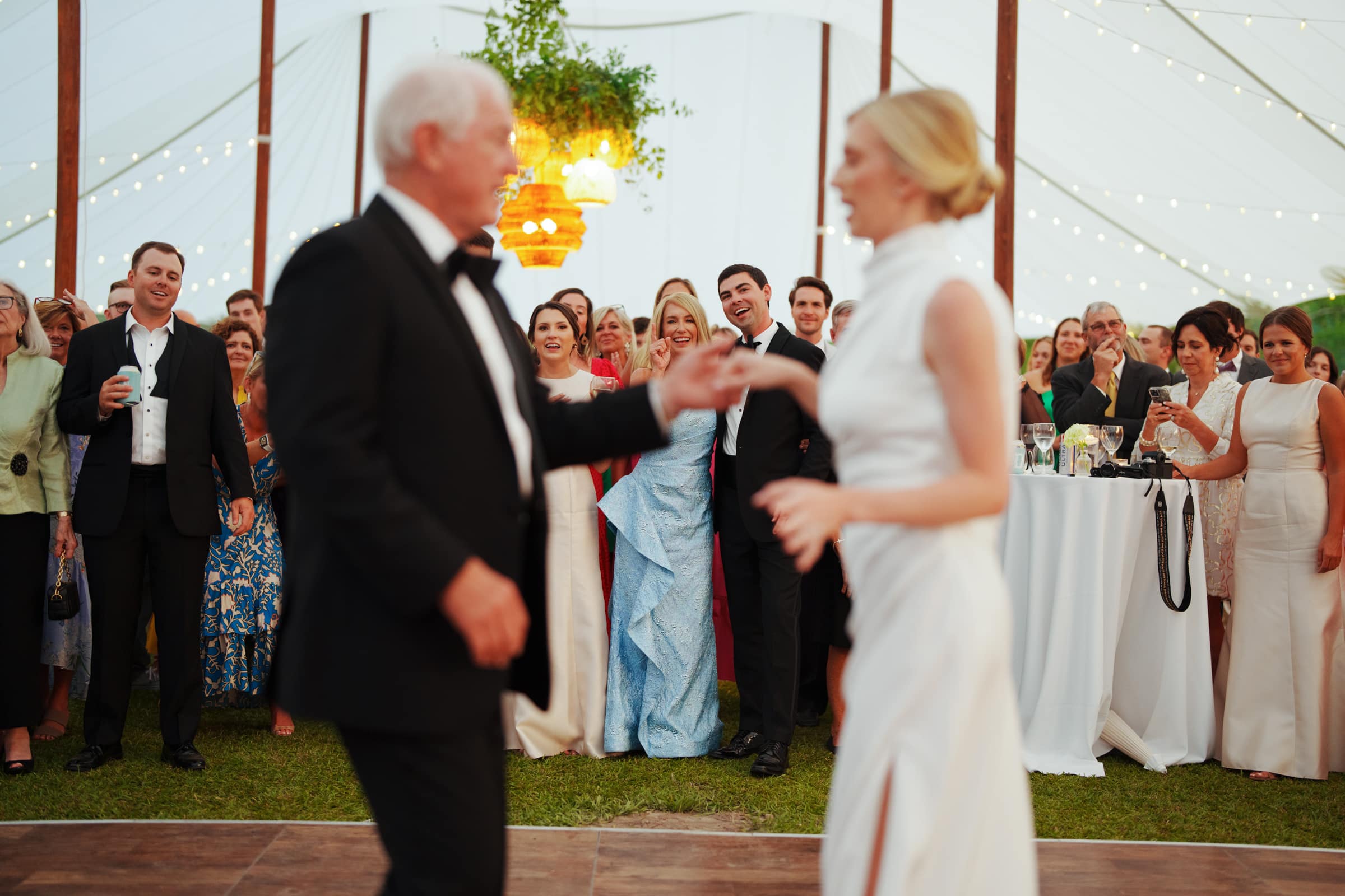 dunn wedding photography - reaves & zach A man in a tuxedo and a woman in a white dress elegantly dance under a tent adorned with string lights, beautifully captured by Dunn wedding photography. Onlookers of various ages and attire watch them, smiling and clapping. The atmosphere is festive and joyful.