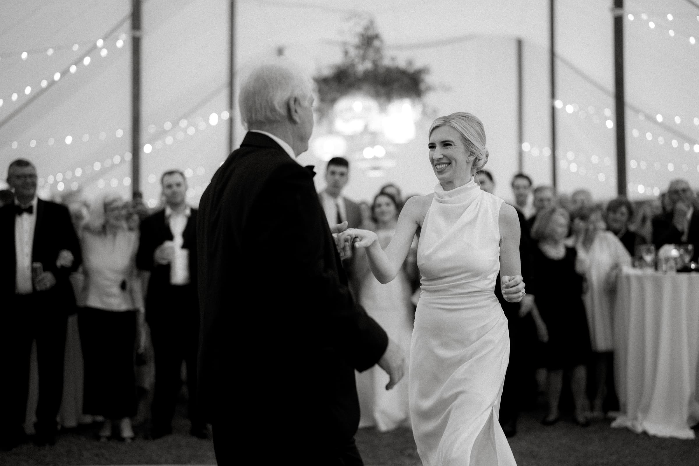 dunn wedding photography - reaves & zach A bride in a white dress dances with an older man in a suit under a tent adorned with string lights. Guests surround them, watching the joyful moment. The black and white image by Dunn wedding photography captures a festive and elegant atmosphere.