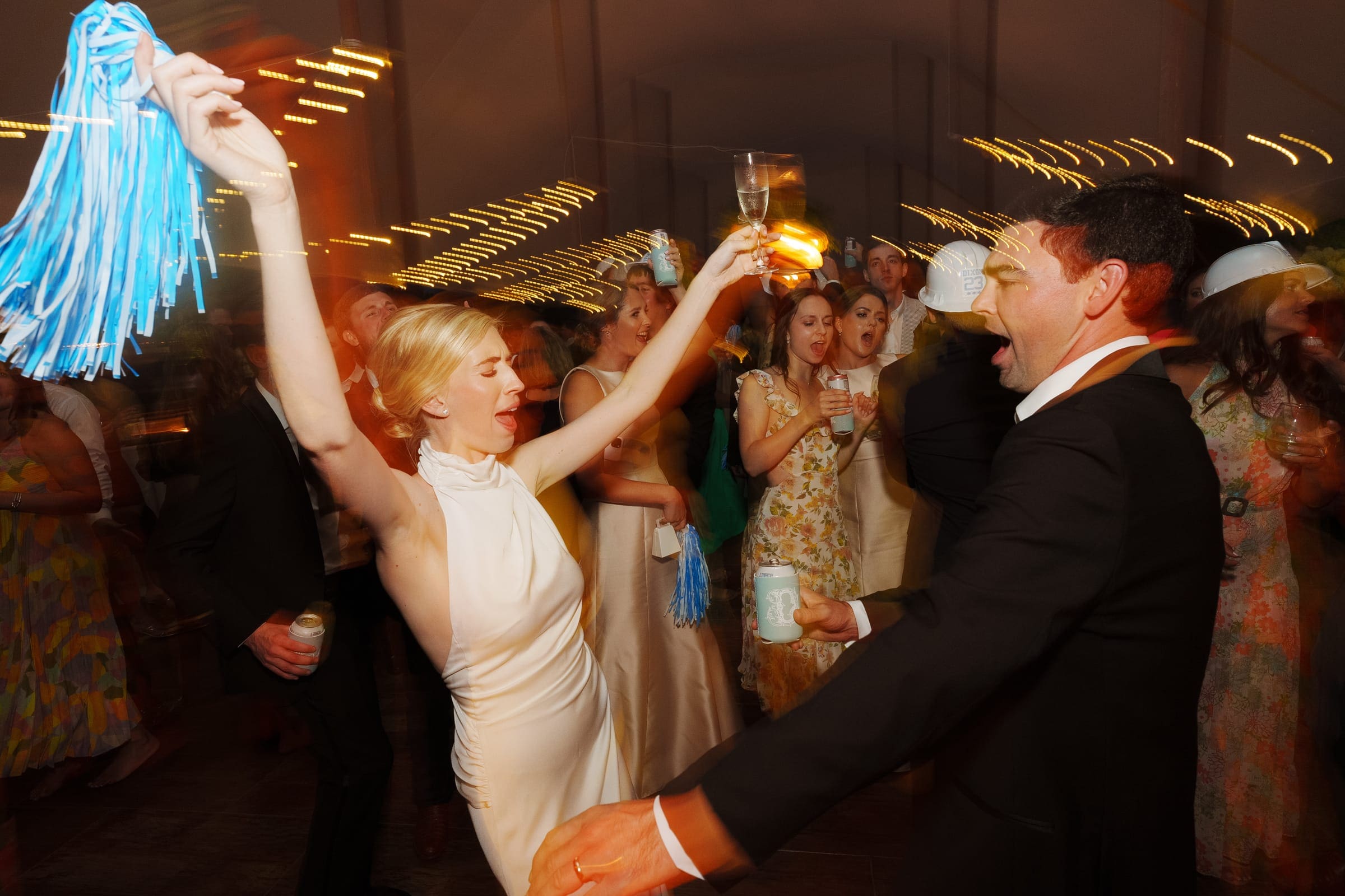 dunn wedding photography - reaves & zach At a festive event captured by Dunn wedding photography, a joyful couple dances energetically. The woman waves a blue pom-pom while holding a drink. Surrounded by others in white hats, everyone savors the lively atmosphere under soft, warm lighting.