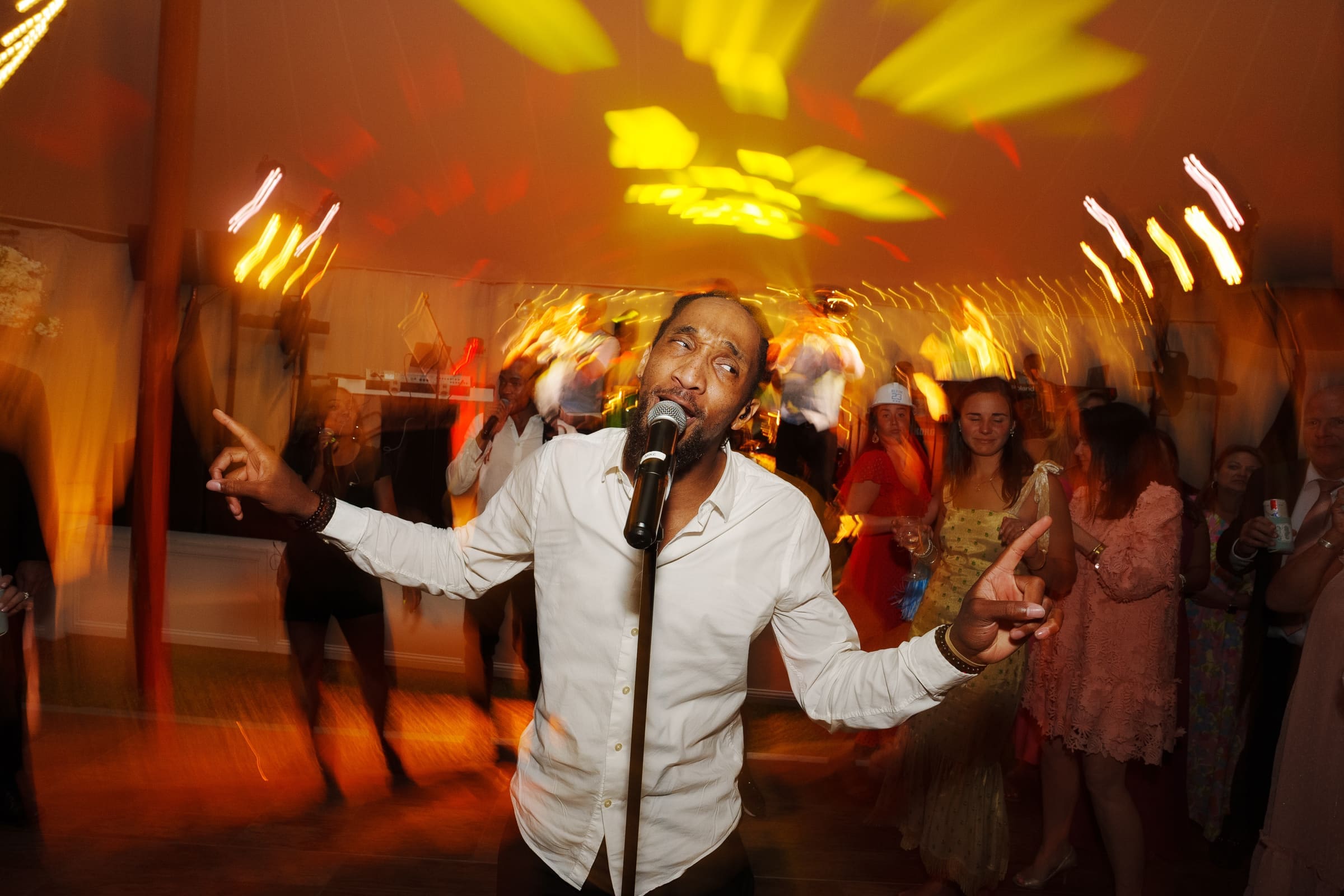 dunn wedding photography - reaves & zach A man in a white shirt is energetically singing into a microphone on a lively dance floor, captured perfectly by Dunn wedding photography. Blurred motion of the colorful lights and surrounding people creates a dynamic, vibrant atmosphere.