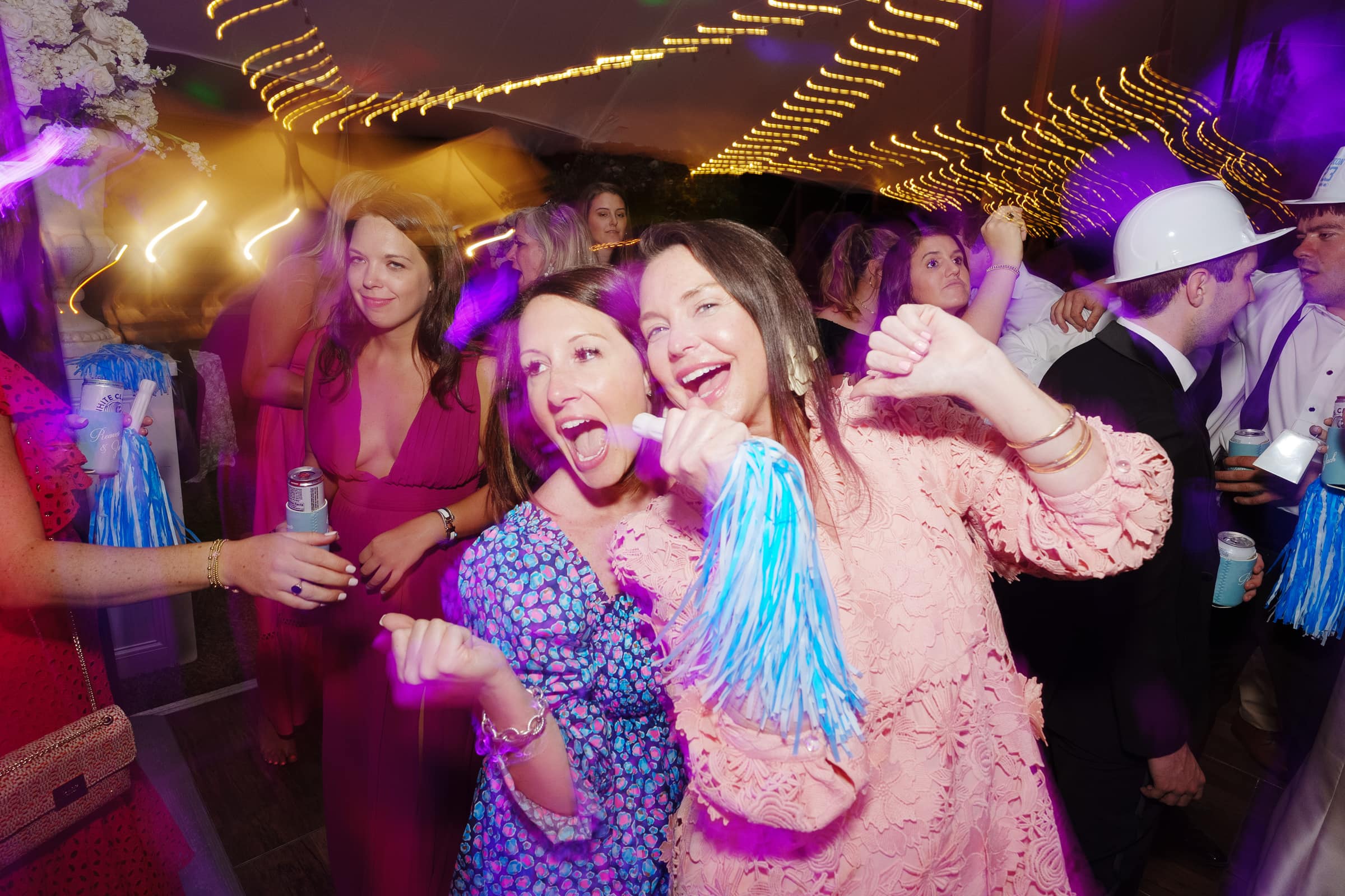 dunn wedding photography - reaves & zach A lively party scene with two smiling women in colorful dresses at the forefront, holding blue pom-poms. String lights by Dunn wedding photography create a vibrant atmosphere, while people in the background enjoy the celebration, some wearing hats and holding drinks.