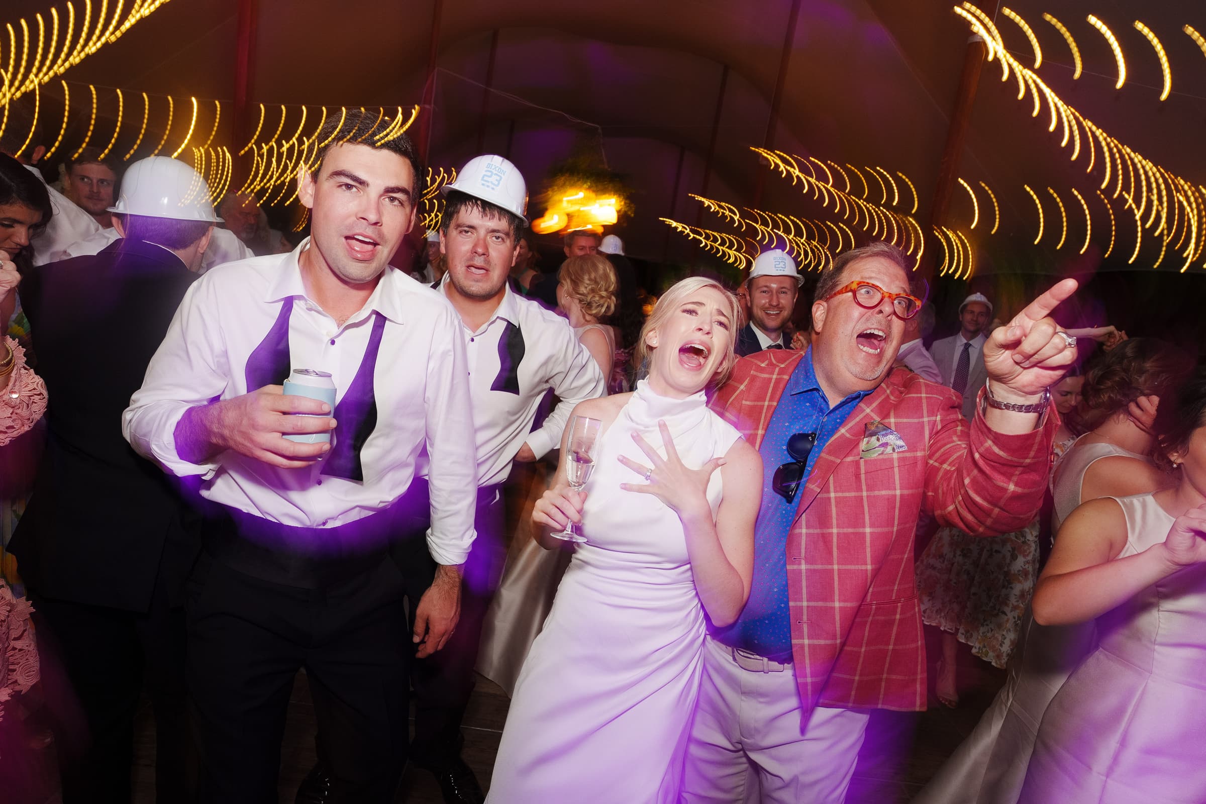 dunn wedding photography - reaves & zach A lively wedding reception captured by Dunn Wedding Photography features four people in the foreground wearing formal attire and sharing drinks. The atmosphere is festive with colorful lighting, as other guests dance energetically in the background.