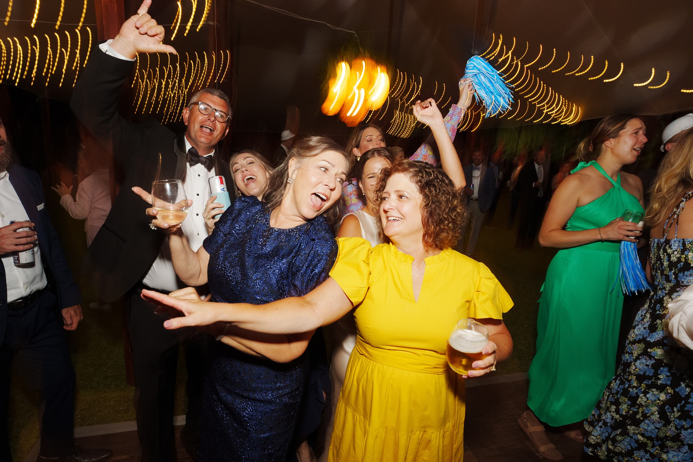dunn wedding photography - reaves & zach A lively group of people dance and celebrate at a party, captured through Dunn wedding photography. The atmosphere is joyful, with women in colorful dresses and a man in a tuxedo holding drinks. Warm string lights create a festive ambiance in the background.